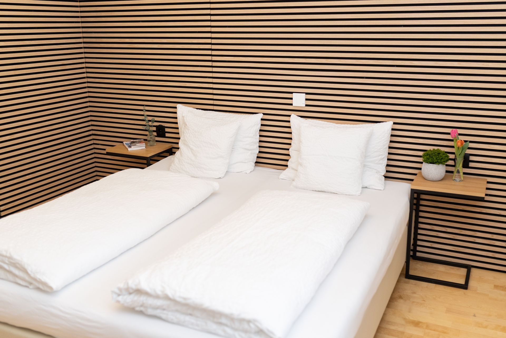 Zimmer Hotel Maria, Doppelbett mit weißen Laken. Kopfteil steht an einer Holzverkleideten Wand.