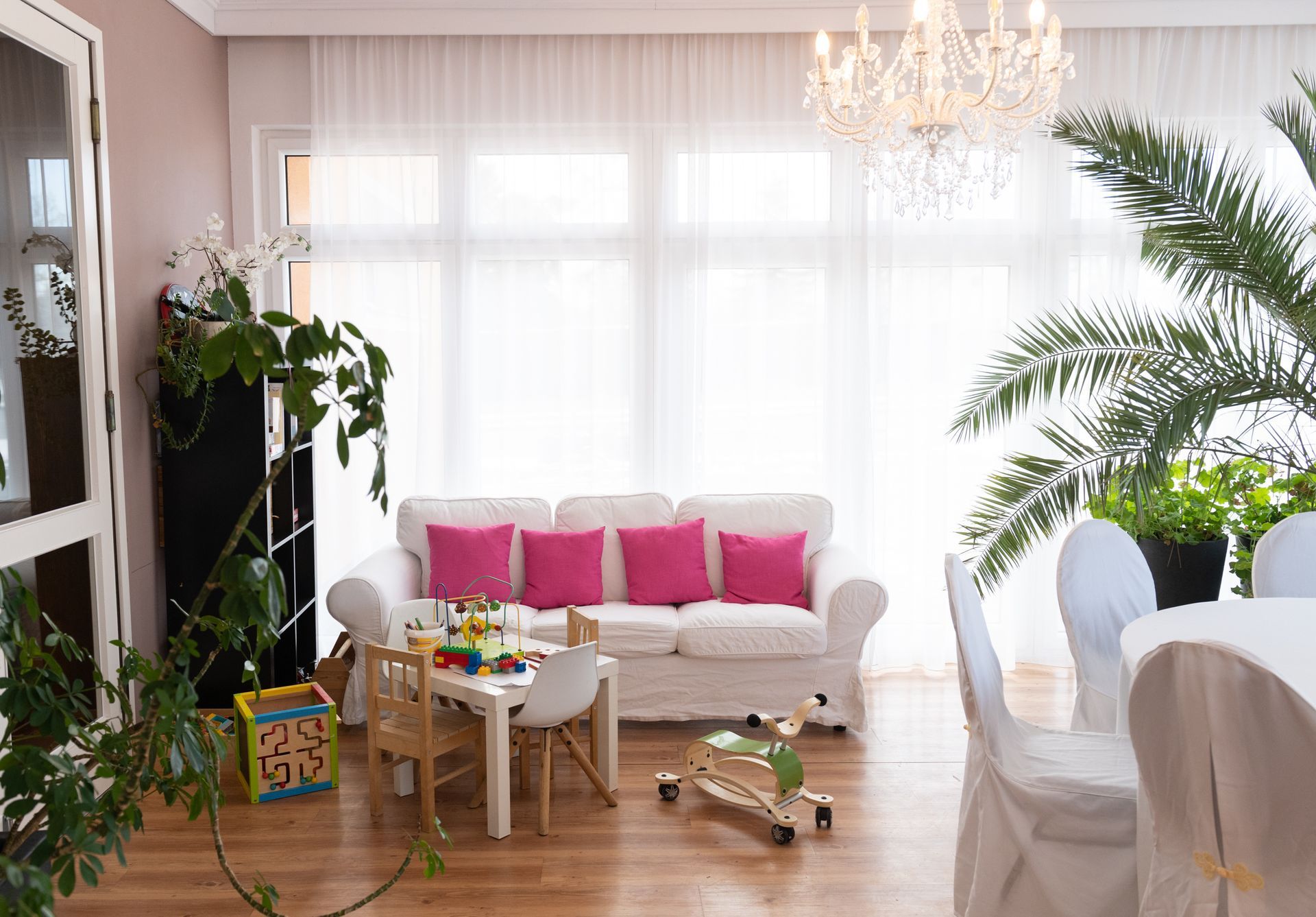 Lounge Hotel Maria, Große Fenster sind mit einem druchsichtigem weißen Vorhang bedeckt. die weiße Couch steht im Fokus, mit pinken Zierkissen. daneben eine große Palme.