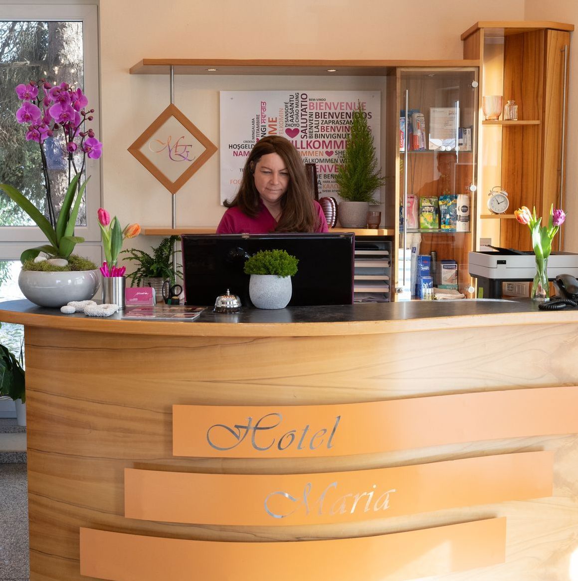 Rezeption von Hotel Maria. Die Rezeption ist aus Holz mit einem Schriftzug an der Front. Auf dem Tisch stehen verschiedene Pflanzen. 