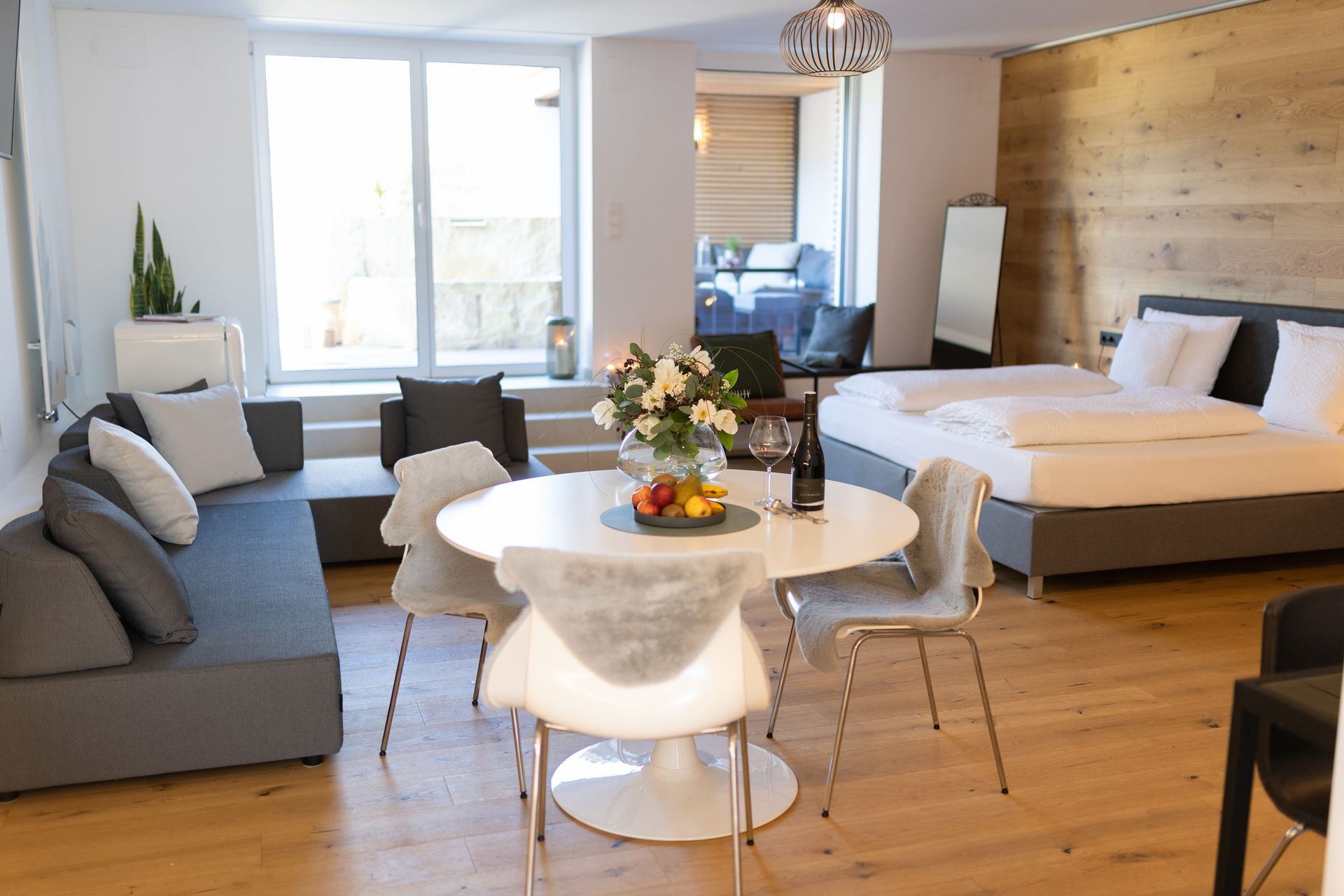Im Fokus ist ein weißer, runder Tisch gedeckt mit Früchten und Wein. vier weiße Stühle rund herum. Im Hintergrund ist das graue Doppelbett mit weißen Laken, sowie ein Großes Fenster und Terrassentüre.