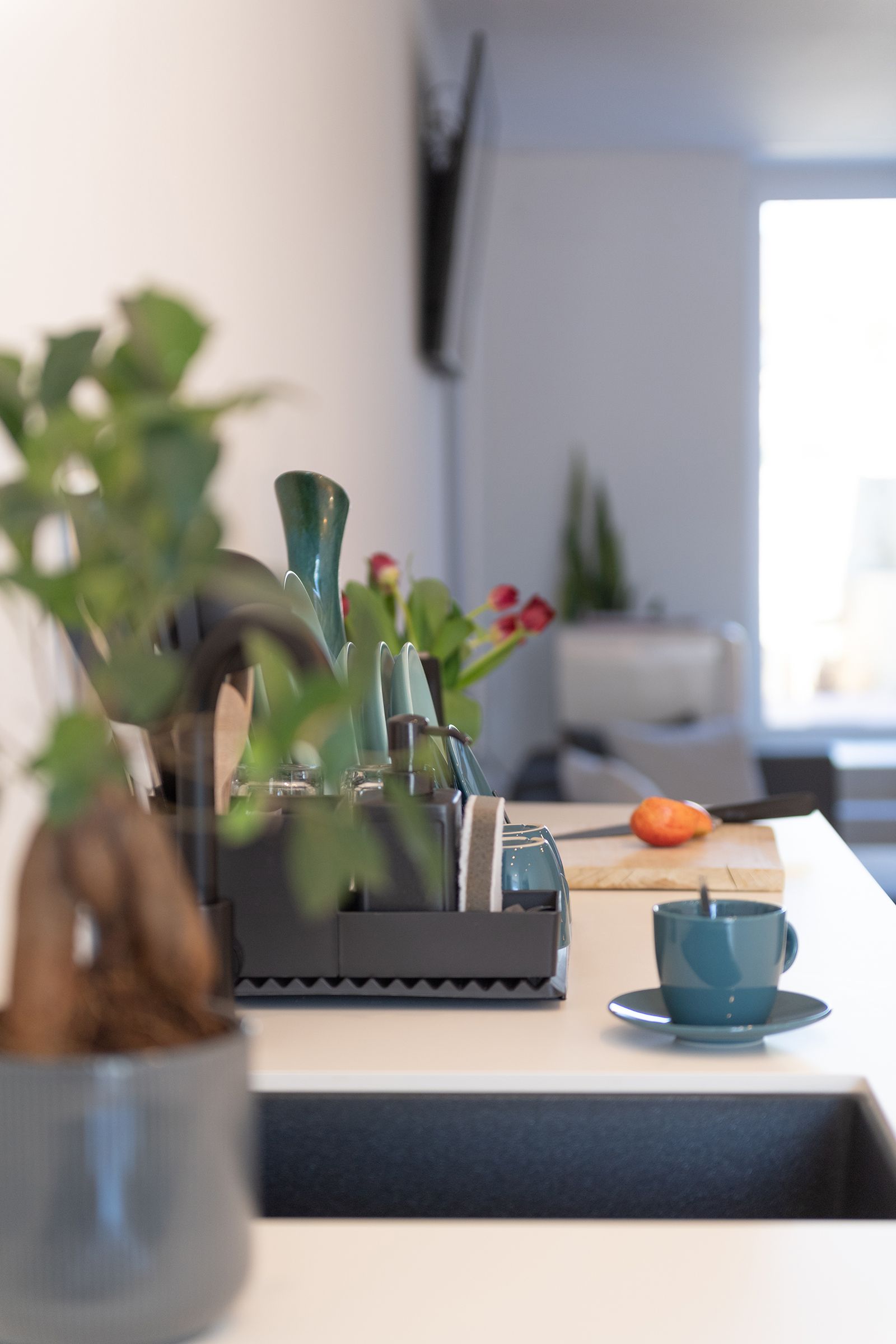 Seitenansicht von weißer Küchenzeile im Fokus. Schwarzes Waschbecken und blaue Teetasse neben schwarzer Kaffeemaschine. Pflanze im Vordergrund nicht im Fokus.
