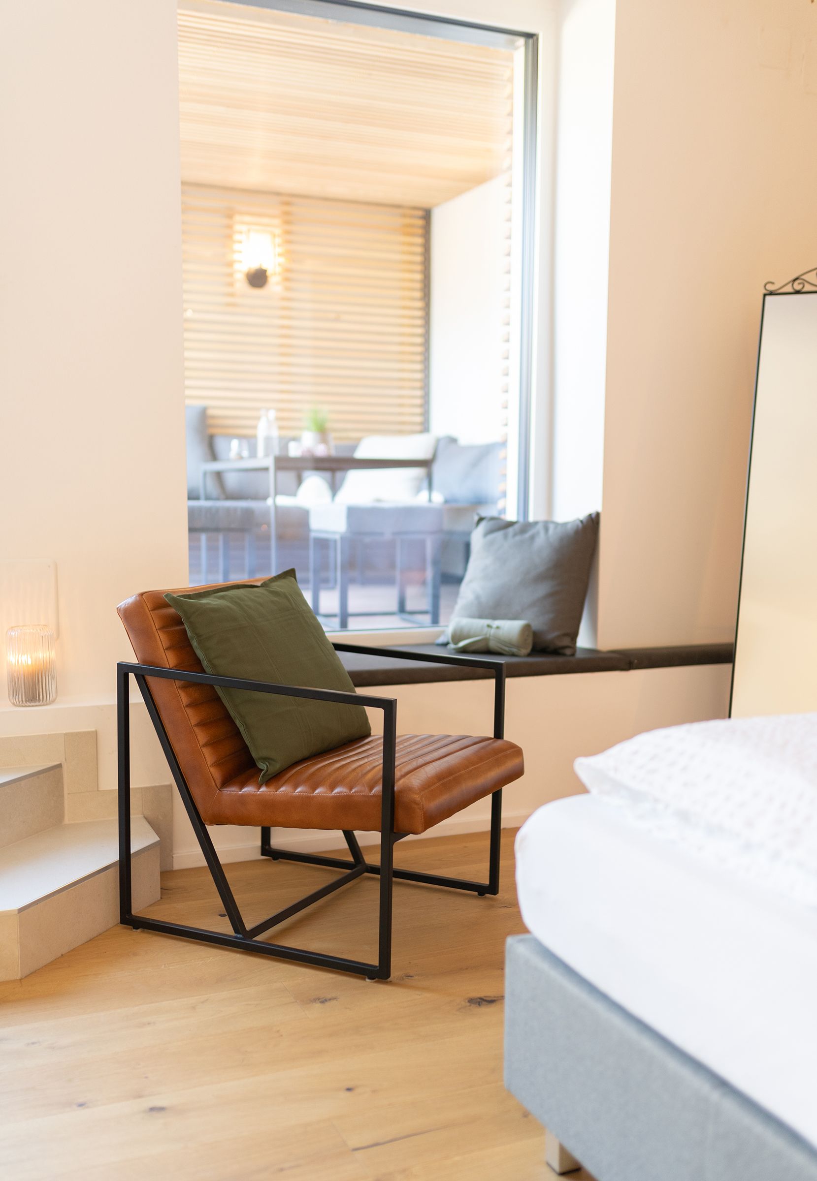 Ein Eck eines Bettes mit weißen Laken. Daneben ein brauner Ledersessel mit schwarzem Metallgestell und einem schwarzen Kissen darauf. Dahinter ein großes Fenster zur Lounge auf der Terrasse.