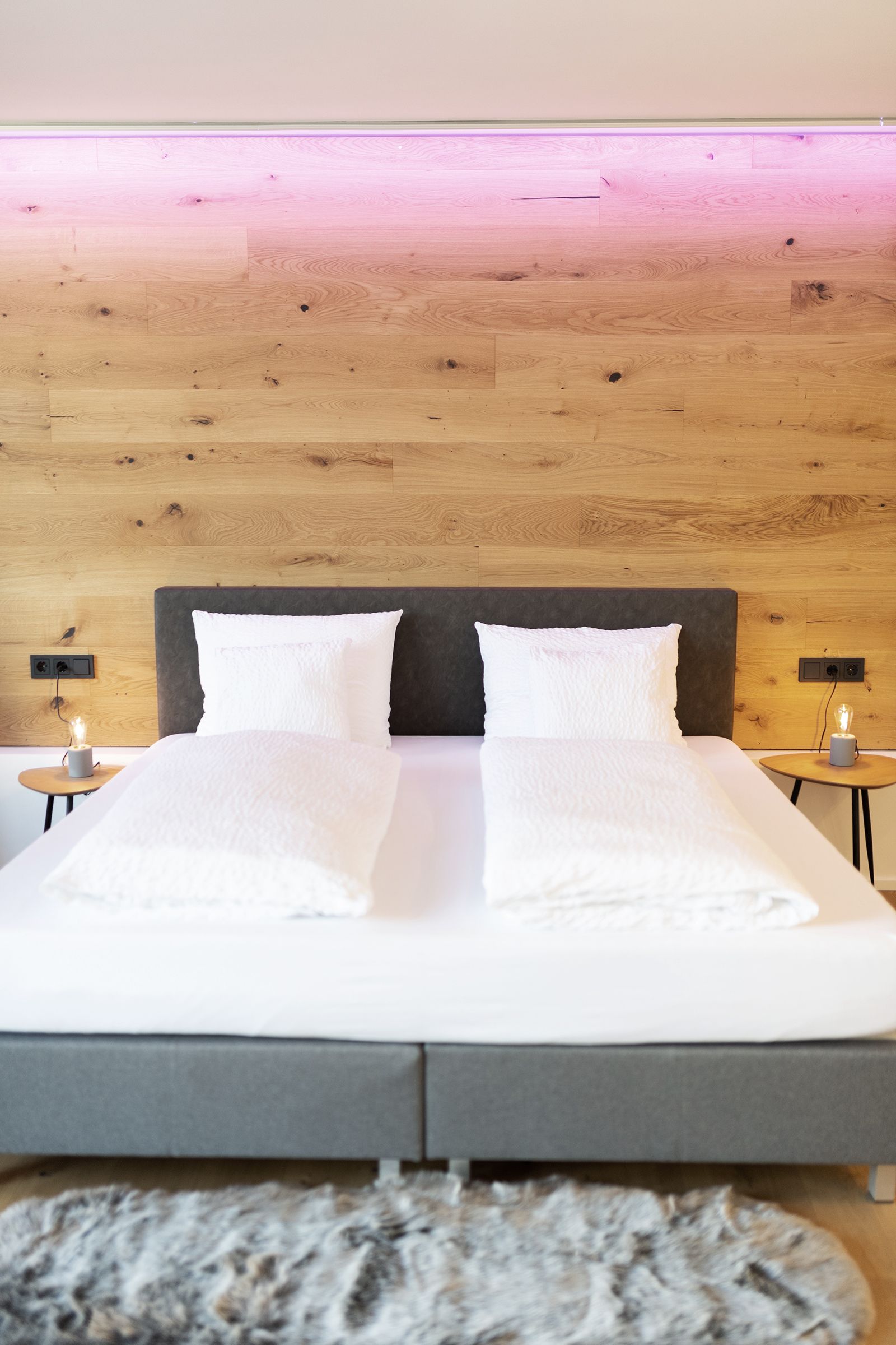 Frontansicht eines Grauen Doppelbettes mit weißen Laken. Das Kopfteil des Bettes steht an einer holzverkleideten Wand mit indirekter rosanen LED Beleuchtung nahe der Decke.