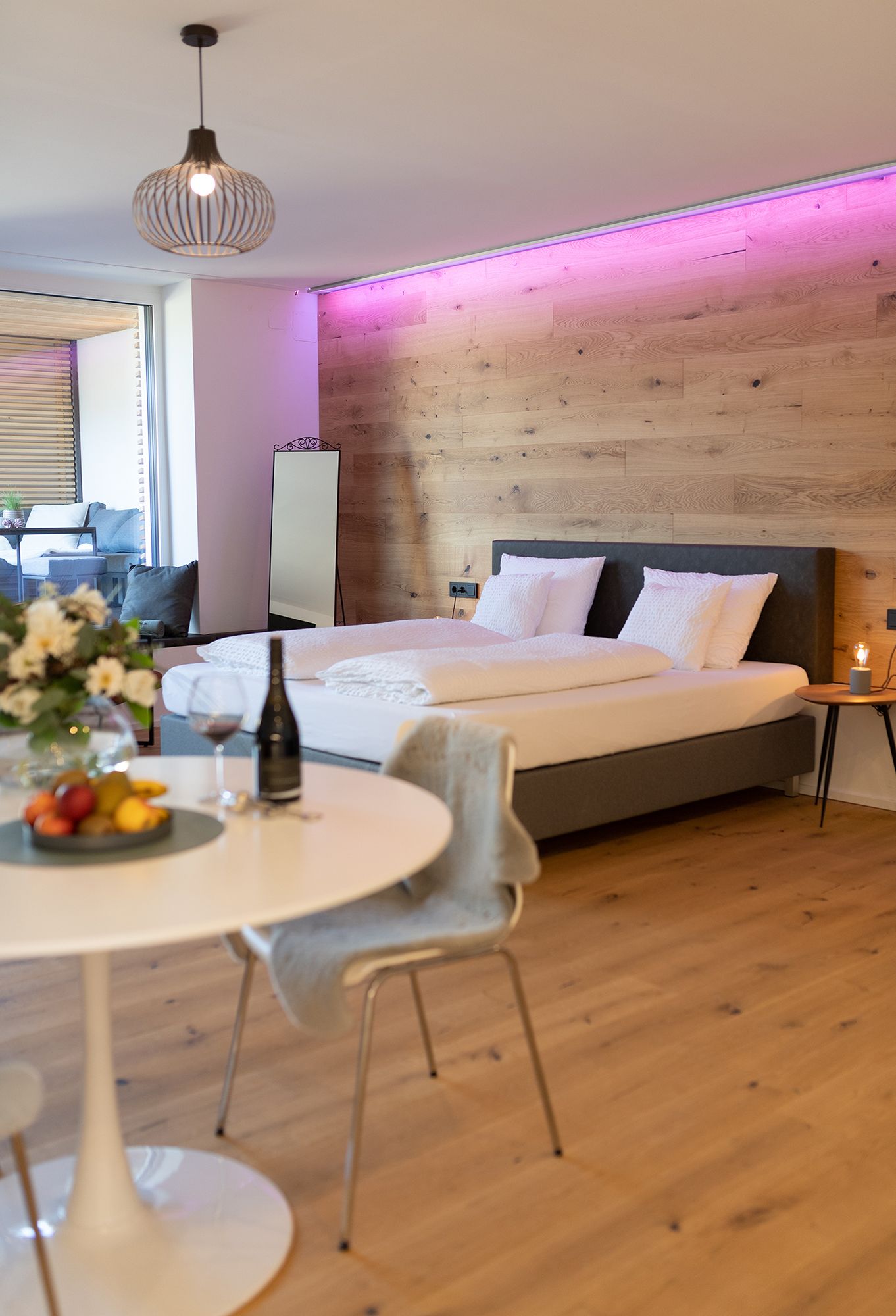 Im Fokus steht das graue Doppelbett mit weißen Laken. Das Bett steht parallel zu einer Holzverkleideten Wand mit indirektem LED Licht nahe der Decke. Im Vordergrund ist ein weißer, runder Tisch gedeckt mit Früchten und Wein.