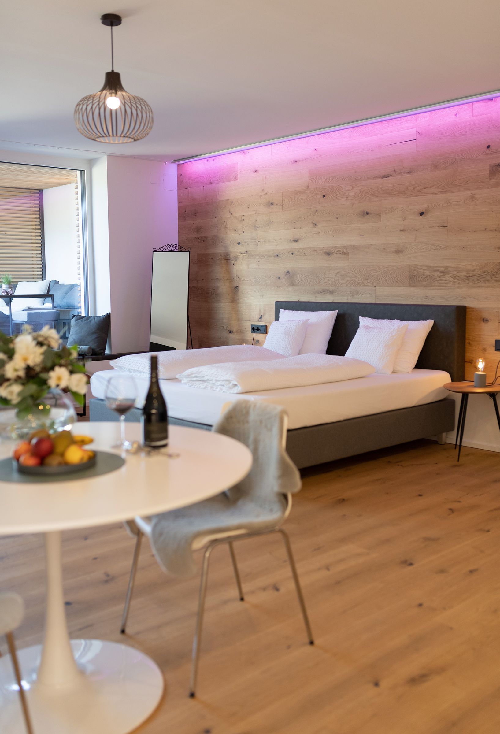 Im Fokus steht das graue Doppelbett mit weißen Laken. Das Bett steht parallel zu einer Holzverkleideten Wand mit indirektem LED Licht nahe der Decke. Im Vordergrund ist ein weißer, runder Tisch gedeckt mit Früchten und Wein.