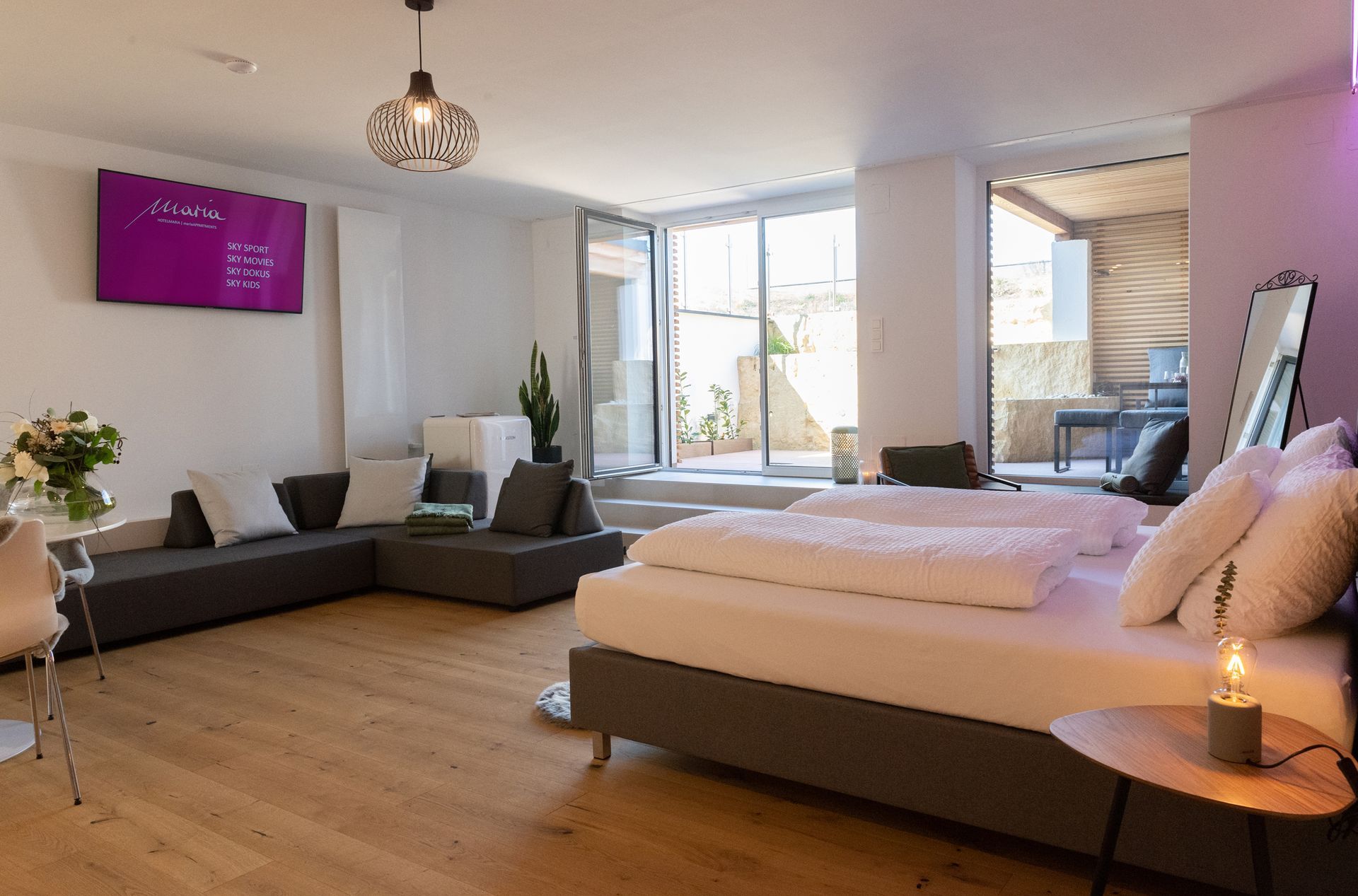 Im Fokus steht das graue Doppelbett mit weißen Laken. Im Hintergrund ist eine graue Eckcouch und darüber ein Fernseher. Schräg neben der Couch ist die große Terassentüre.