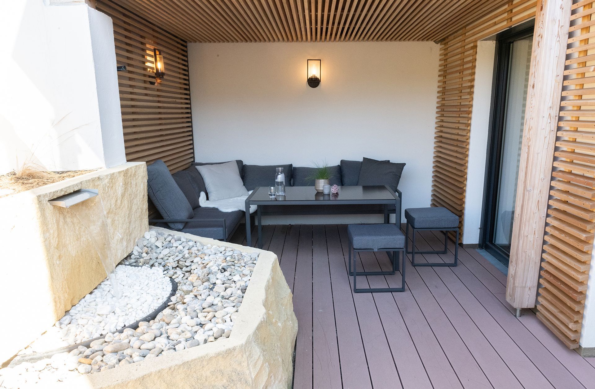 Die Lounge auf der Terrasse besteht aus einer grauen Eckcouch, einem schwarzen Tisch und zwei Hocker im Stil der Couch. Ein Teil des Steinbrunnen ist am Rand des Bildes zu sehen.