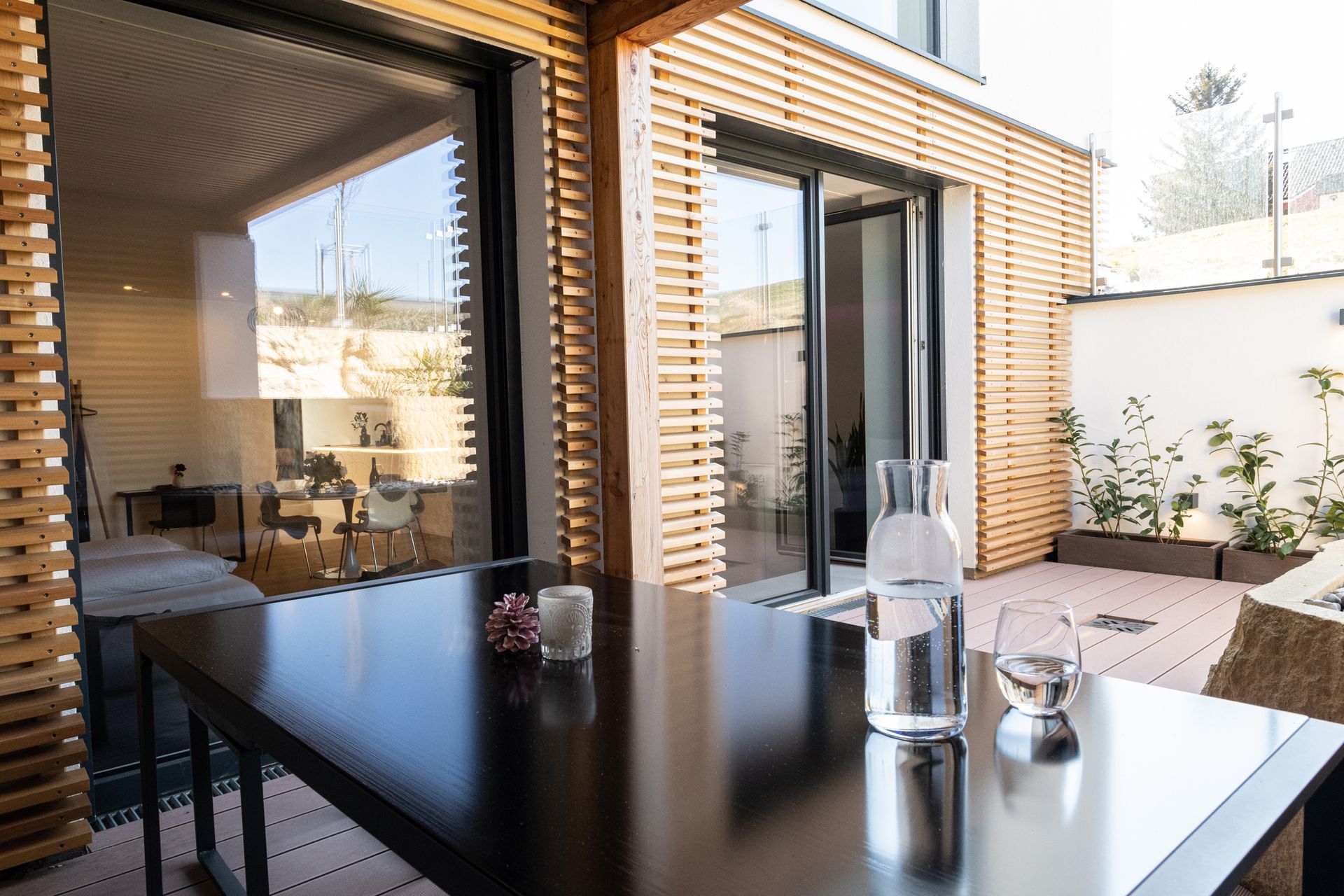 Ansicht auf die holzverkleidete Außenwand der Terrasse, über den schwarzen Tisch hinweg. Auf dem Tisch steht eine Wasserkaraffe aus glas und ein volles Glas daneben.