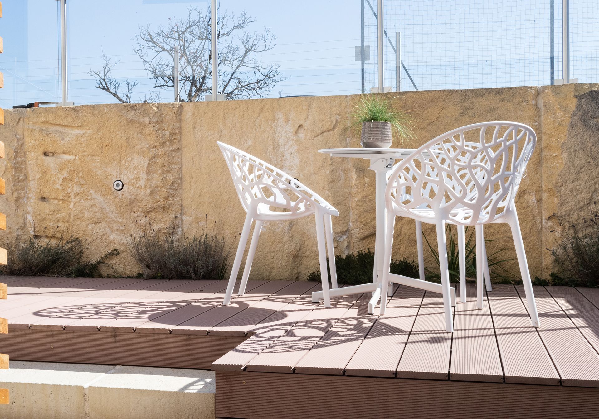 auf einer höhergelegten Terrasse mit Holzboden steht ein weißer runder Tisch und zwei weiße Stühle. Im Hintergrund ist eine beige Wand und der Himmel zu sehen.