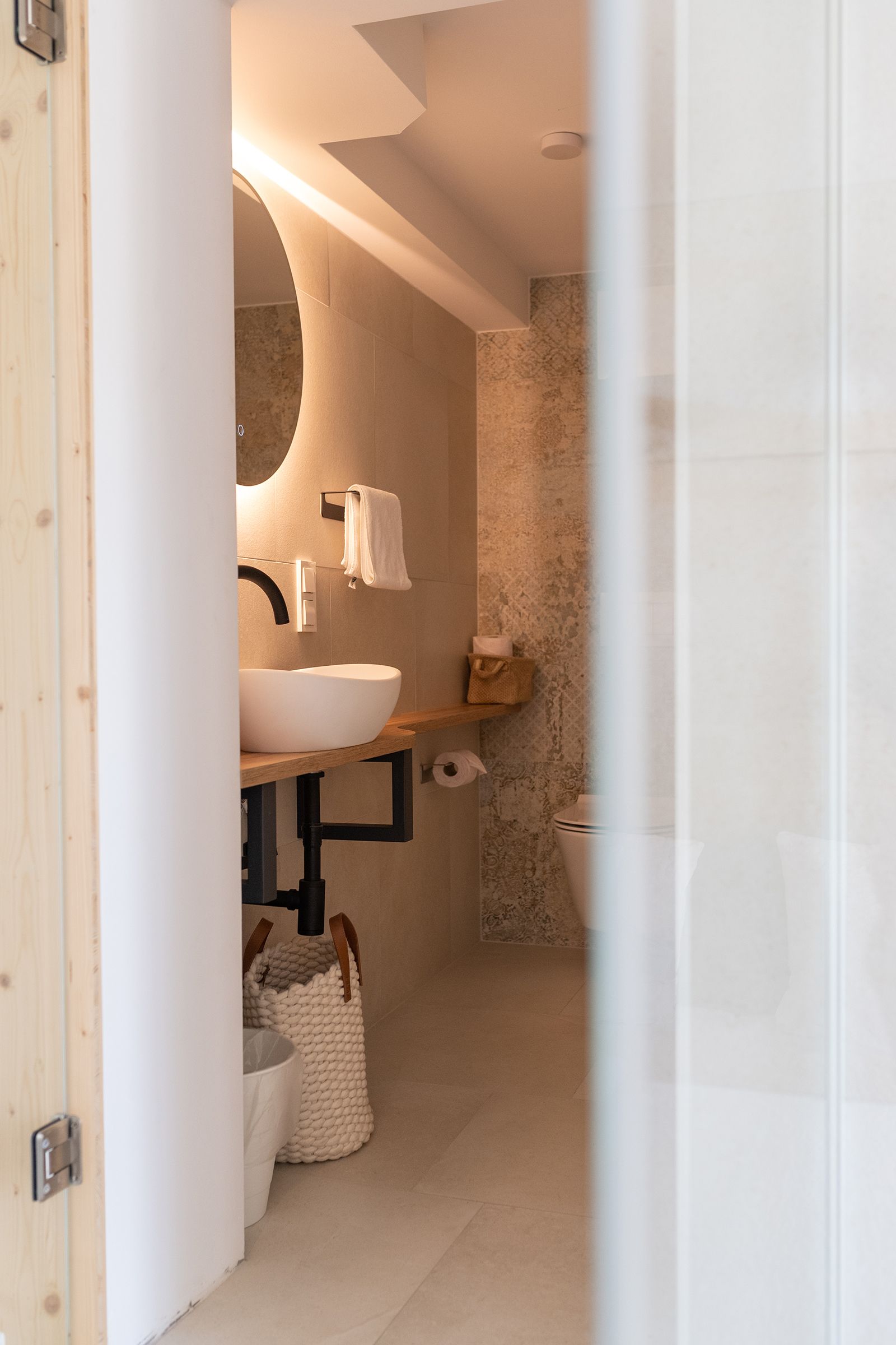 Blick in das Badezimmer durch eine Türe hindurch. Warme indirekte Beleuchtung nahe der Decke. Weißes Waschbecken auf Holztisch. Schwarze Armaturen und Rohre aus Metall. Über dem Waschbecken hängt ein großer runder Spiegel.