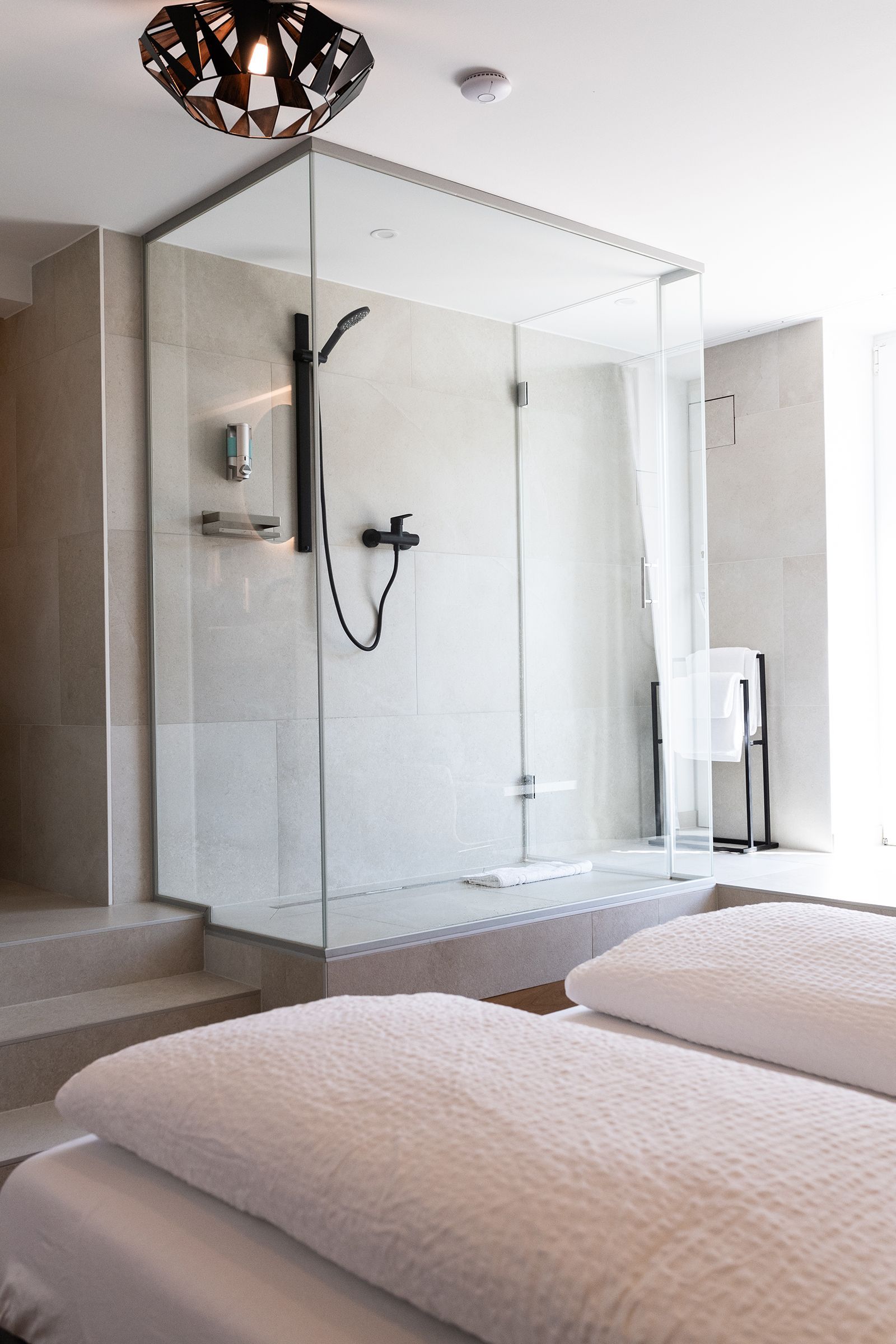 Gesamte Glasdusche ist zusehen, mit schwarzen Armaturen und Handtuchhalter daneben. Der Eingang ist schräg gegenüber. Die hälfte des grauen Bettes mit weißen Laken in rechter unteren Ecke des Bildes.