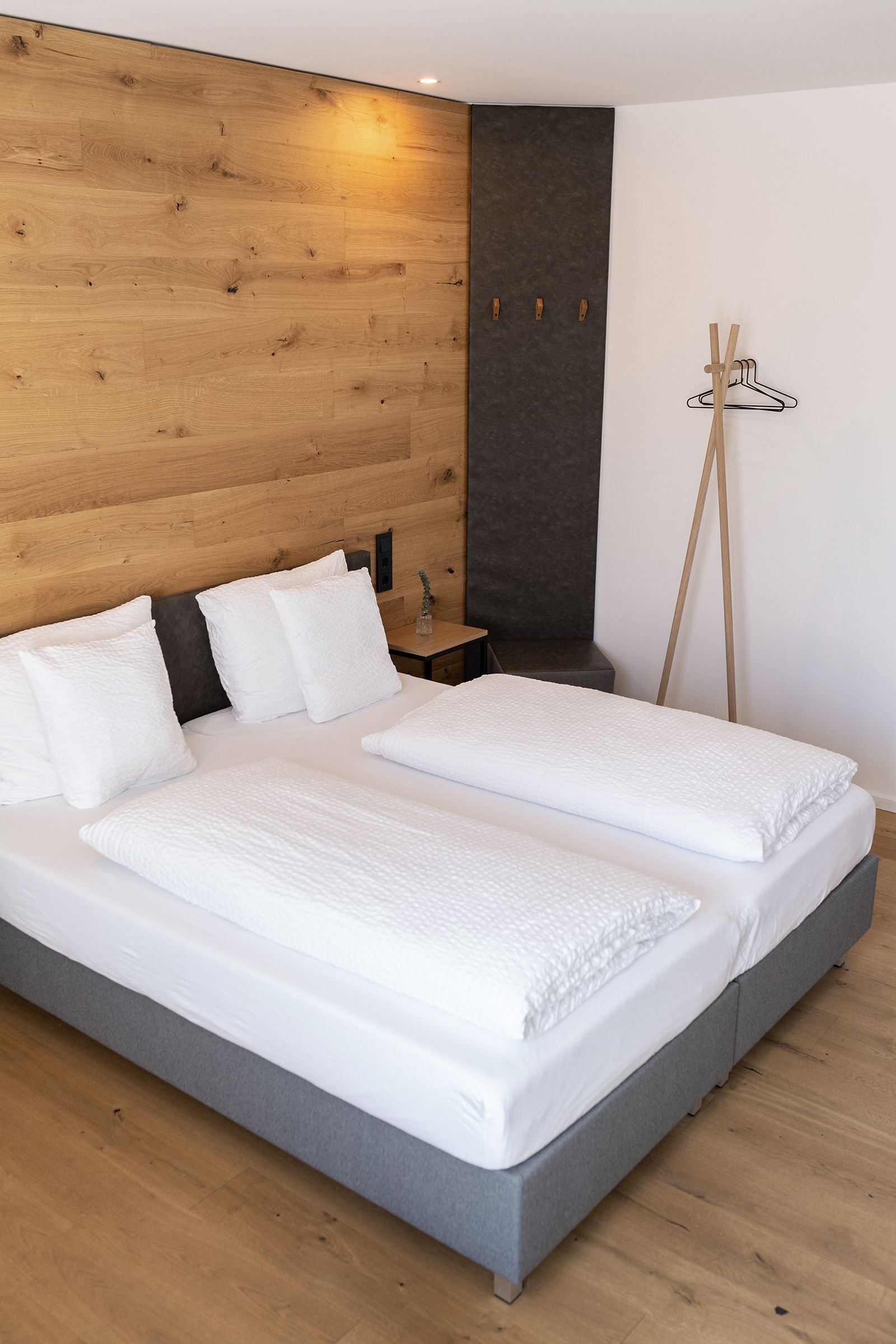 Graues Doppel-Boxspringbett mit weißen Laken an einer holzverkleideten Wand. Daneben ein kleiner Nachttisch mit Pflanze. Daneben kleine Holzgarderobe.