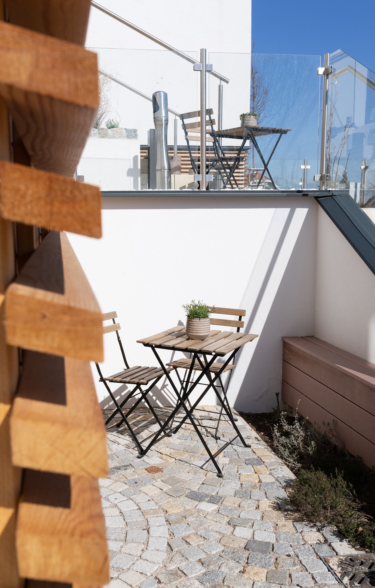 Halbhohe Betonwand. Davor steht ein kleiner Holztisch mit passendem Sessel. Auf den Tisch ist eine kleine, grüne Pflanze. Die Terrasse ist Pflasterstein.  Blauer Himmel im Hintergrund.