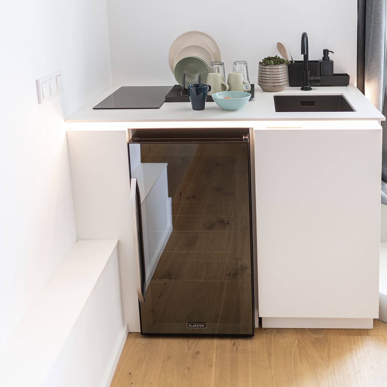 Kleine, weiße Küchenzeile mit schwarzen Armaturen und Kühlschrank. Darauf befindet sich verschieden-farbenes Geschirr, links daneben ist ein schwarzer Induktionsherd und rechts daneben ist ein schwarzes Waschbecken.