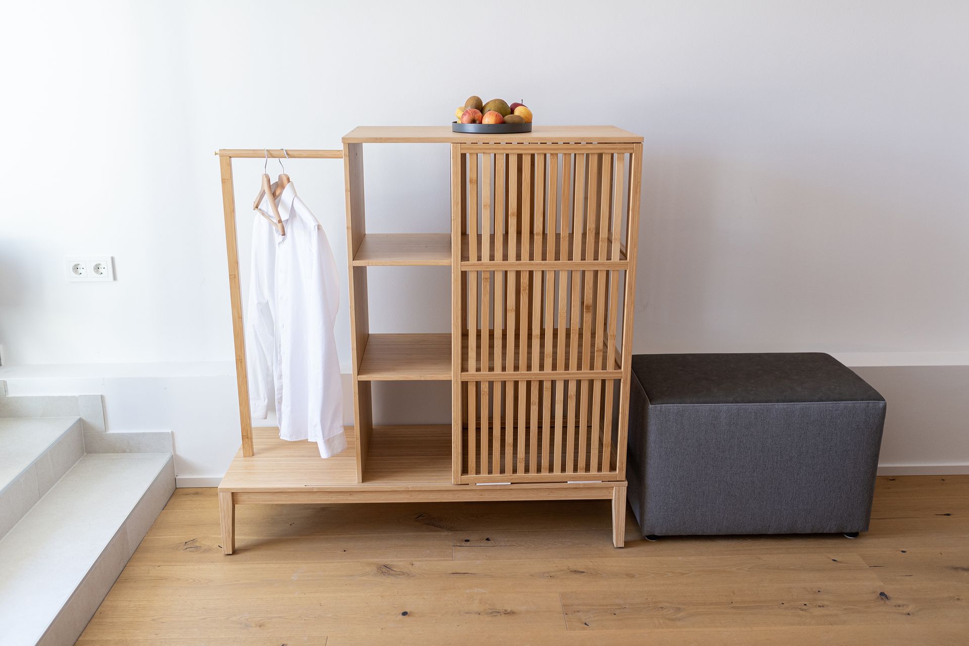 Stilvoll eingerichteter Eingangsbereich mit einer Holzgarderobe und kleinem offenen Schrank. Auf dem Regal steht eine Schale mit frischem Obst, daneben hängt ein weißes Hemd auf Holzkleiderbügeln. Ein grauer Hocker ergänzt das minimalistische, helle Interieur mit Holzfußboden und klaren Linien.