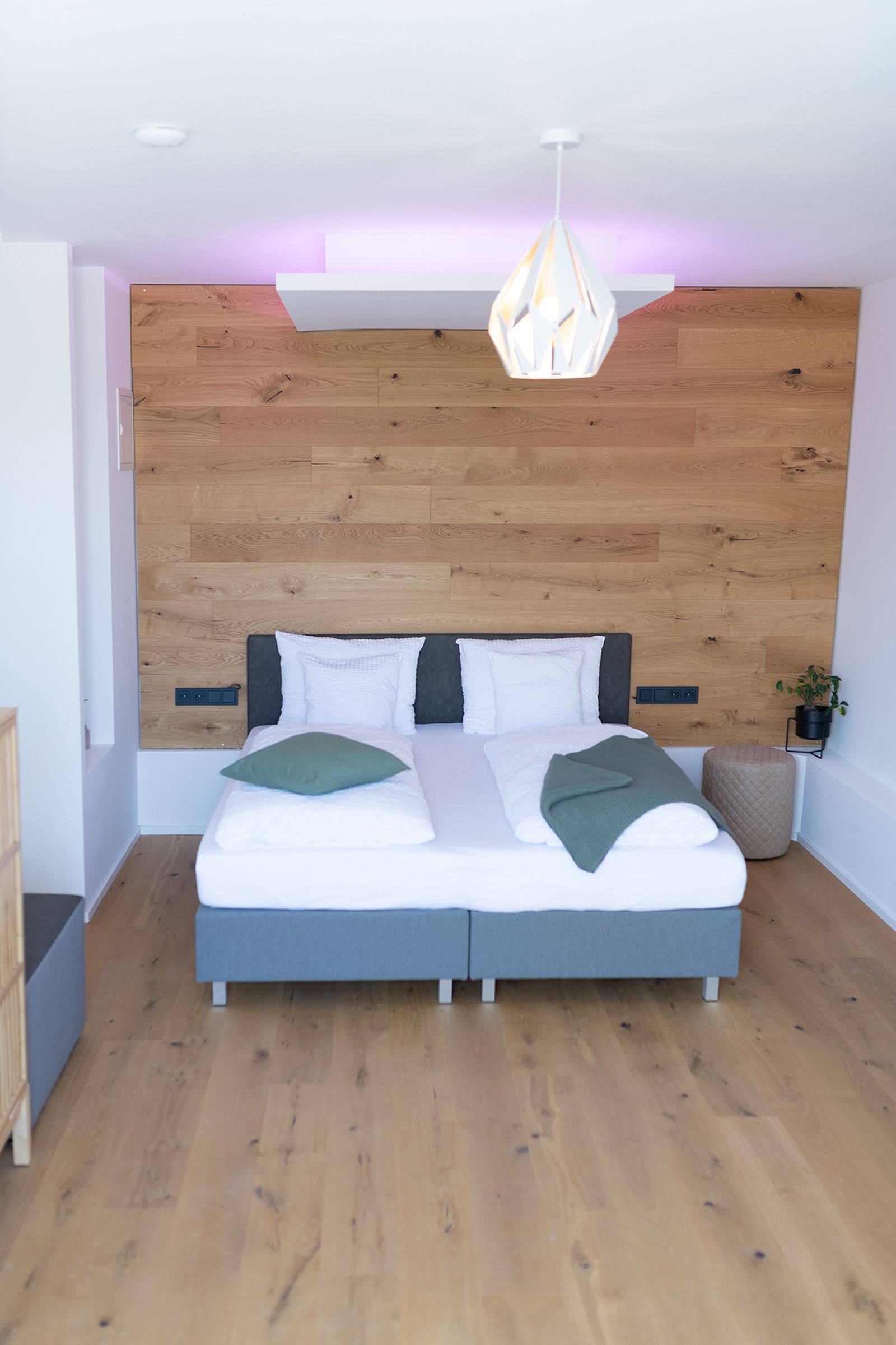 graues Doppelbett steht im Fokus. Der Kopfteil des Bettes steht an einer Holzverkleideten Wand. Der Boden ist ebenso aus Parkett. Rechts neben dem Bett steht ein brauner Hocker und eine Pflanze.