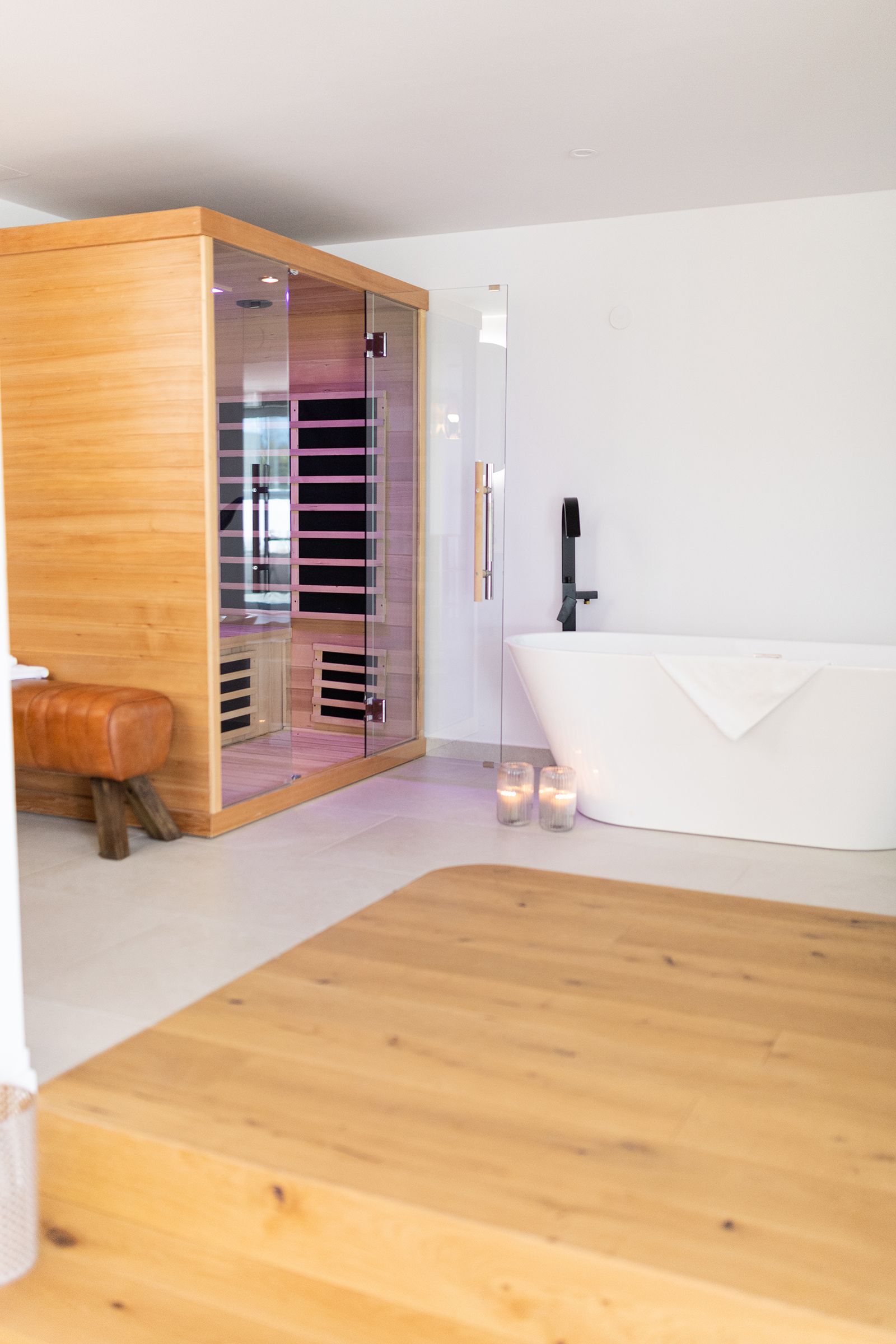 Freistehende Badewanne mit schwarzer Armatur und zwei Kerzengläsern am Wannenrand. Daneben befindet sich eine stilvolle Infrarotsauna mit Holzverkleidung und Glasfront. Daneben ist ein brauner Lederhocker. Der Raum ist mit teils hellem Parkett und weißen Wänden gestaltet.