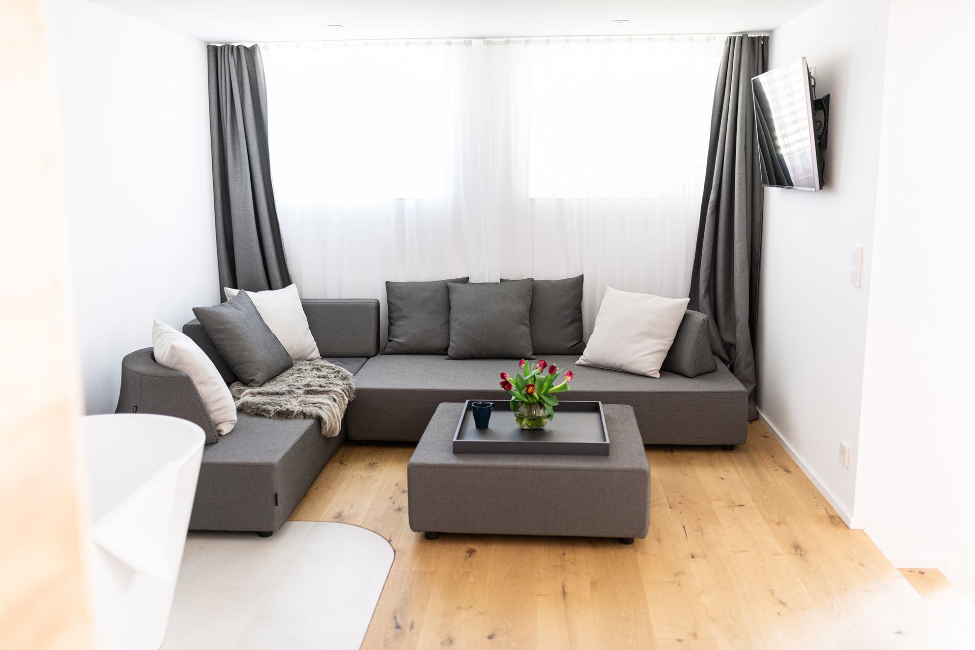 Die graue Couch steht im Fokus, darüber zwei Fenster mit weißem Vorhang. Auf dem Couchtisch ist das Tablett mit roten Blumen. Schräg daneben ist ein Teil der weißen, freistehenden Badewanne zu sehen. 