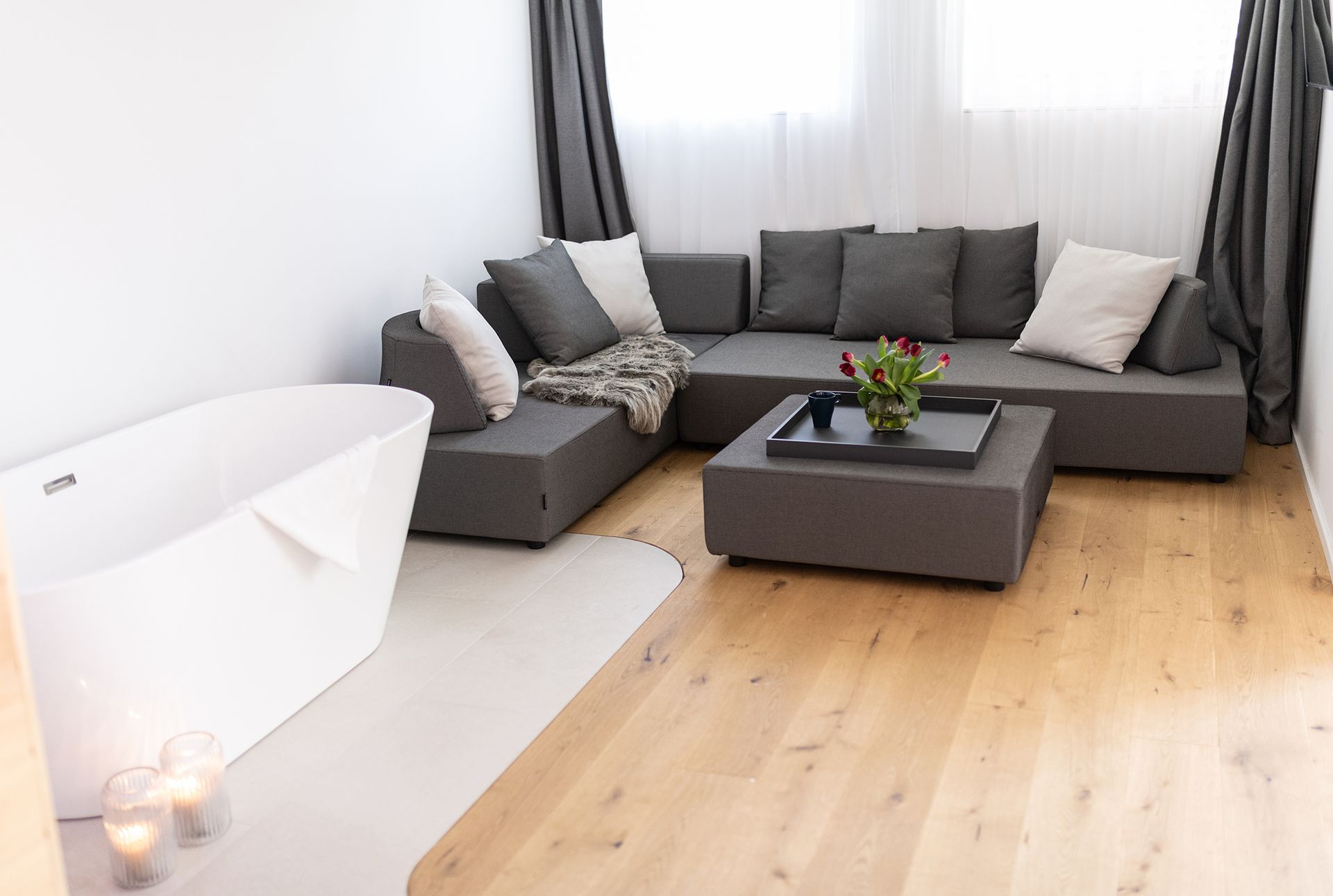Die graue Couch steht im Fokus, darüber zwei Fenster mit weißem Vorhang. Auf dem Couchtisch ist das Tablett mit roten Blumen. Schräg daneben ist ein Teil der weißen, freistehenden Badewanne zu sehen. 