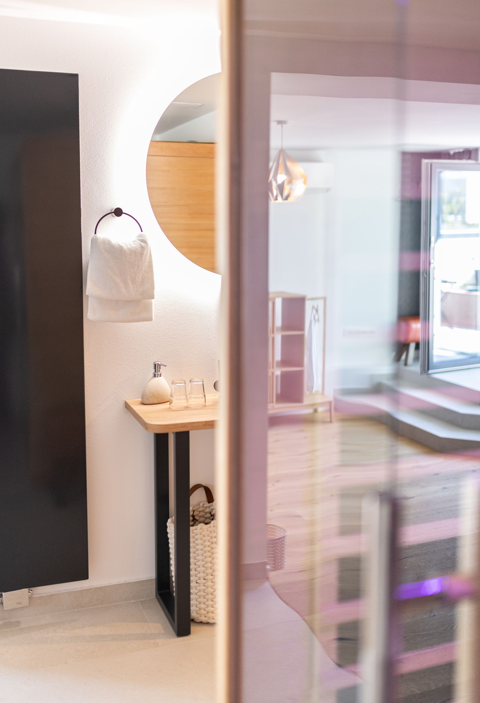 Badezimmer hinter Infrarotkabine. Weißes Waschbecken auf Holztisch mit schwarzen Armaturen und Leitungen. Ein großer runder Spiegel hängt über dem Waschbecken. Indirekte, helle Beleuchtung hinter dem Spiegel.