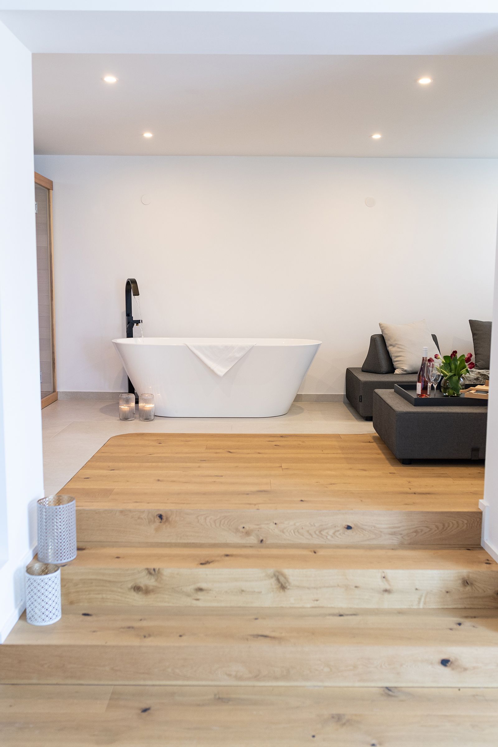 Holzparkettstufen führen zu einer freistehenden, weißen Badewanne mit schwarzen Armaturen. Rechts davon ist ein Teil der grauen Couch zu sehen. Ein Tablett mit Blumen und Wein ist darauf angerichtet. 