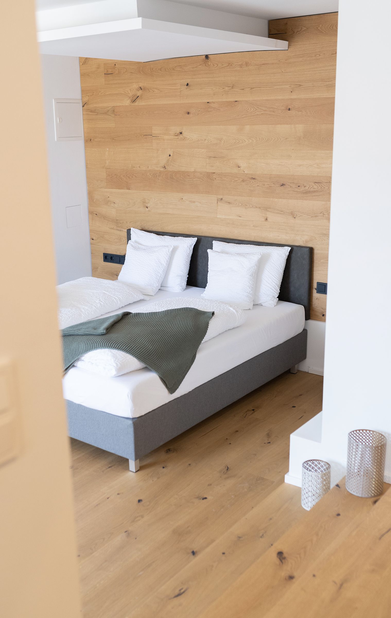 Das graue doppel-Boxspringbett mit weißen Laken steht im Fokus. Der Boden ist Parkett. Das Bett steht mit Kopfteil zu einer holzverkleideten Wand. 