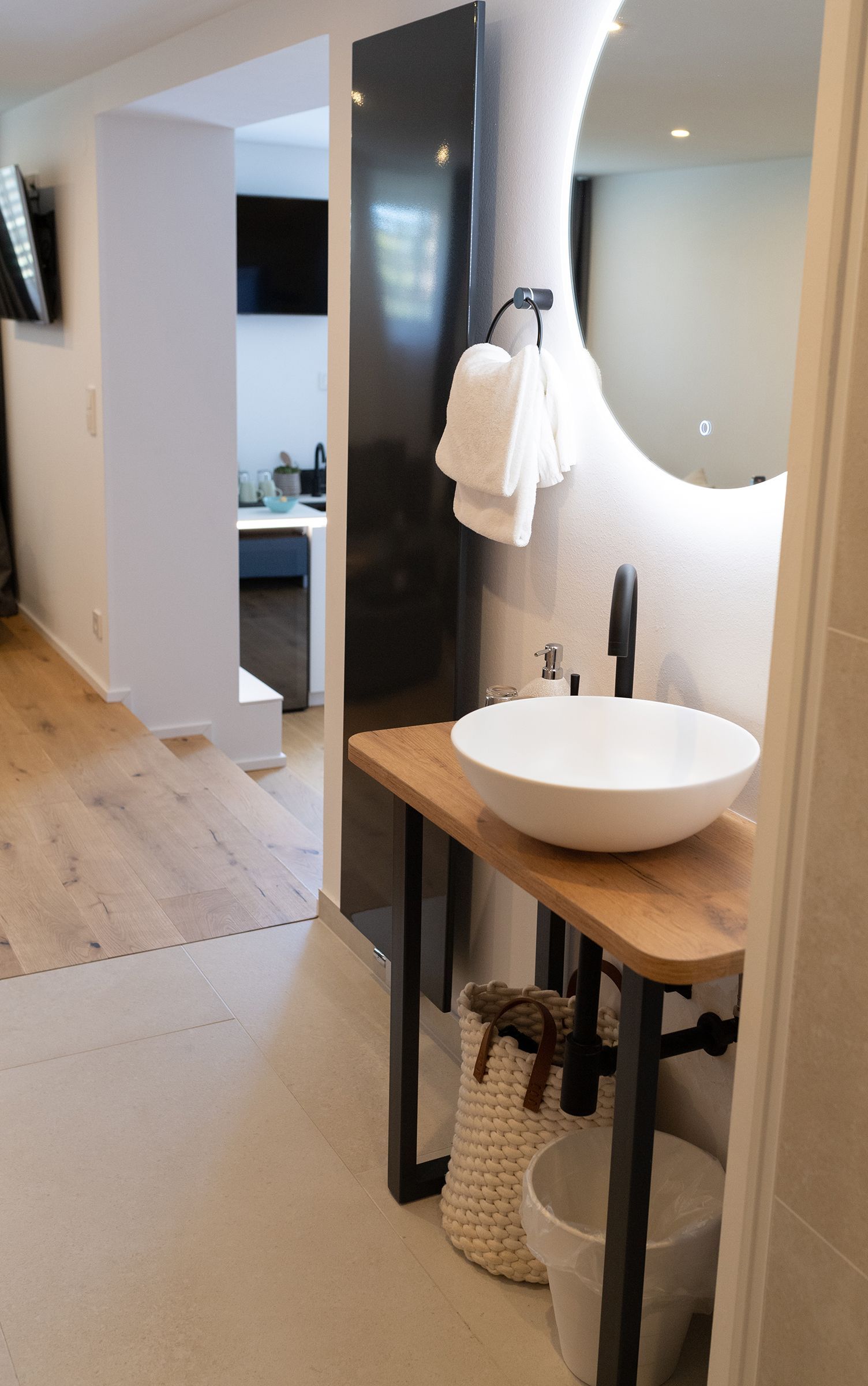 Weißes Waschbecken auf Holztisch mit schwarzen Armaturen und Leitungen. Ein großer runder Spiegel hängt über dem Waschbecken. Indirekte, helle Beleuchtung hinter dem Spiegel. 