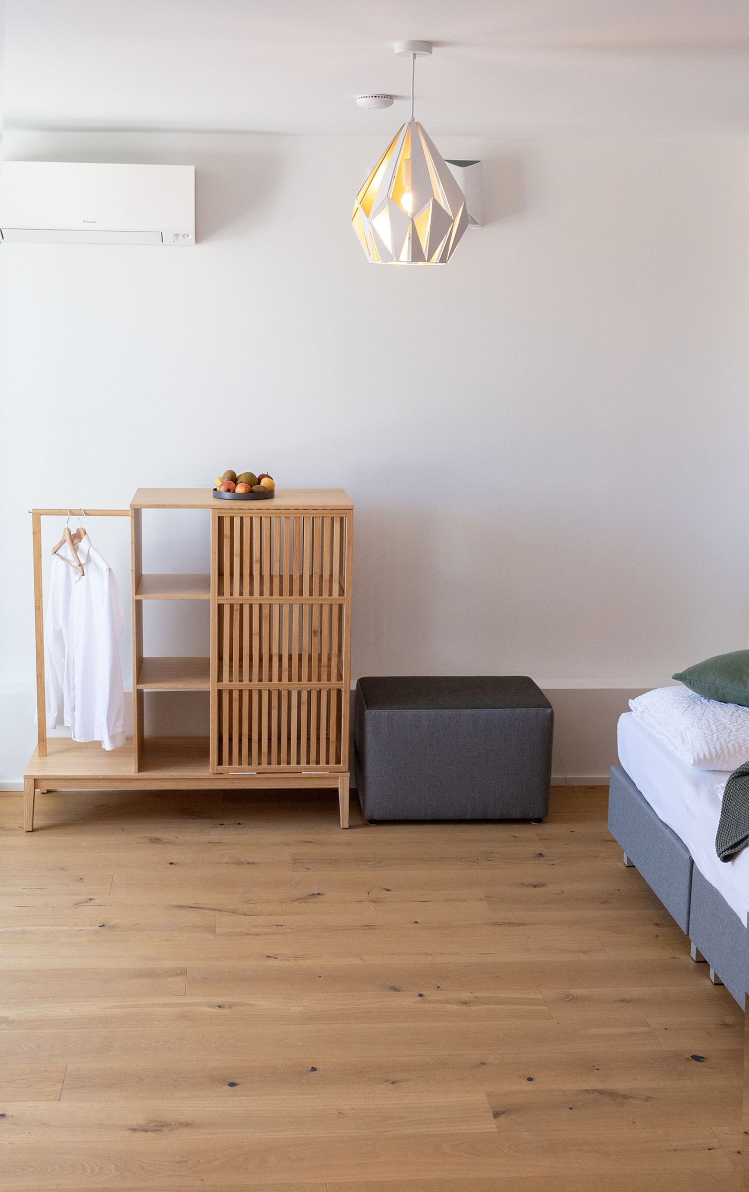 Stilvoll eingerichteter Eingangsbereich mit einer Holzgarderobe und kleinem offenen Schrank. Auf dem Regal steht eine Schale mit frischem Obst, daneben hängt ein weißes Hemd auf Holzkleiderbügeln. Ein grauer Hocker ergänzt das minimalistische, helle Interieur mit Holzfußboden.