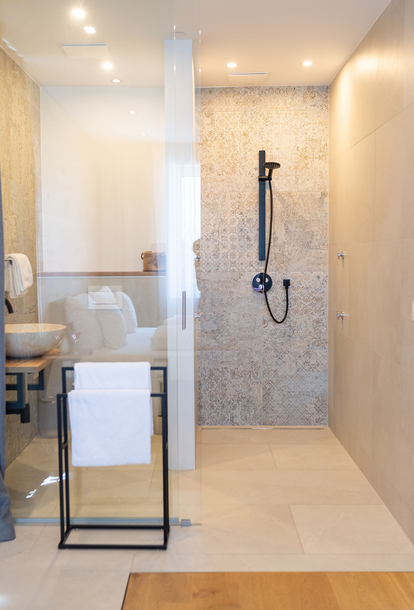 Moderne begehbare Dusche mit großer Handbrause und schwarzer Armatur. Die Rückwand ist mit strukturierten, beige-grauen Fliesen im Vintage-Dekor gestaltet, die Seitenwände und der Boden in hellen Naturtönen gehalten. Mit Glas abgetrennt ist daneben ein modernes Waschbecken und WC