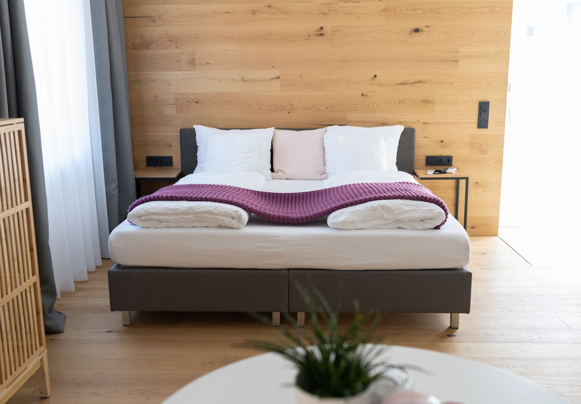 graues Doppelbett mit weißen Laken steht im Fokus. Der Kopfteil des Bettes steht an einer Holzverkleideten Wand. Der Boden ist ebenso aus Parkett. Rechts und links neben dem Bett stehen Nachttische. Im Vordergrund ist ein weißer runder Tisch zu sehen mit einer Pflanze darauf.