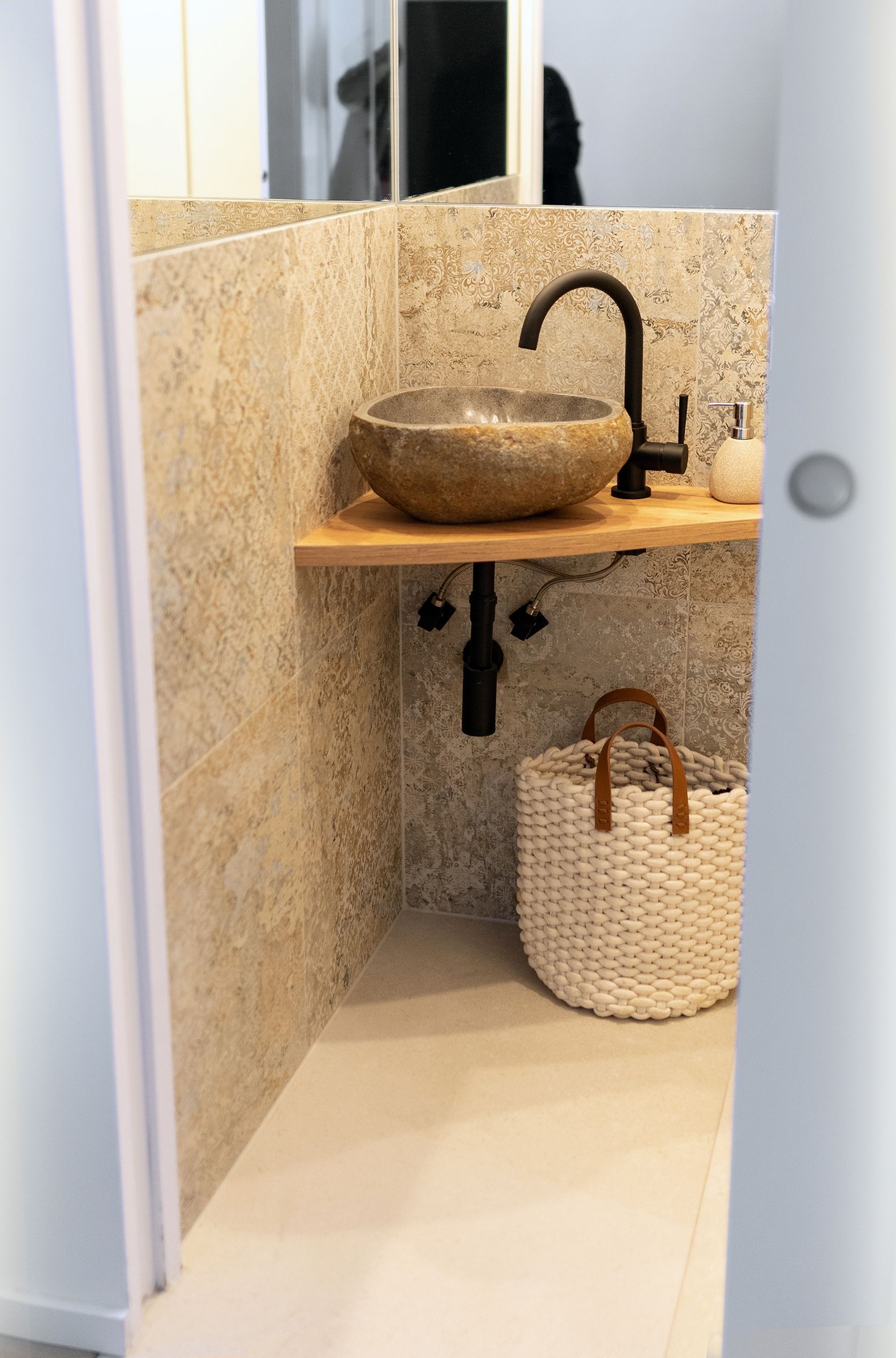 Ein rundes Stein Waschbecken auf einem Holztisch mit schwarzer Armatur. darüber ist ein Spiegel. Die Rückwand ist mit strukturierten, beige-grauen Fliesen im Vintage-Dekor gestaltet. Unter dem Waschtisch ist ein Korb.