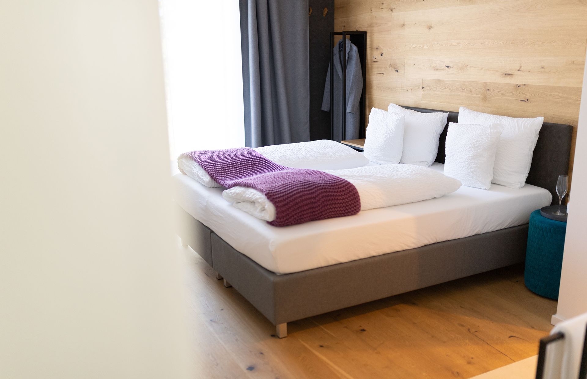 Das graue doppel-Boxspringbett mit weißen Laken steht im Fokus. Der Boden ist Parkett. Das Bett steht mit Kopfteil zu einer holzverkleideten Wand. Neben dem Bett ist eine große Fentstertüre mit Verdunklungsvorhängen.