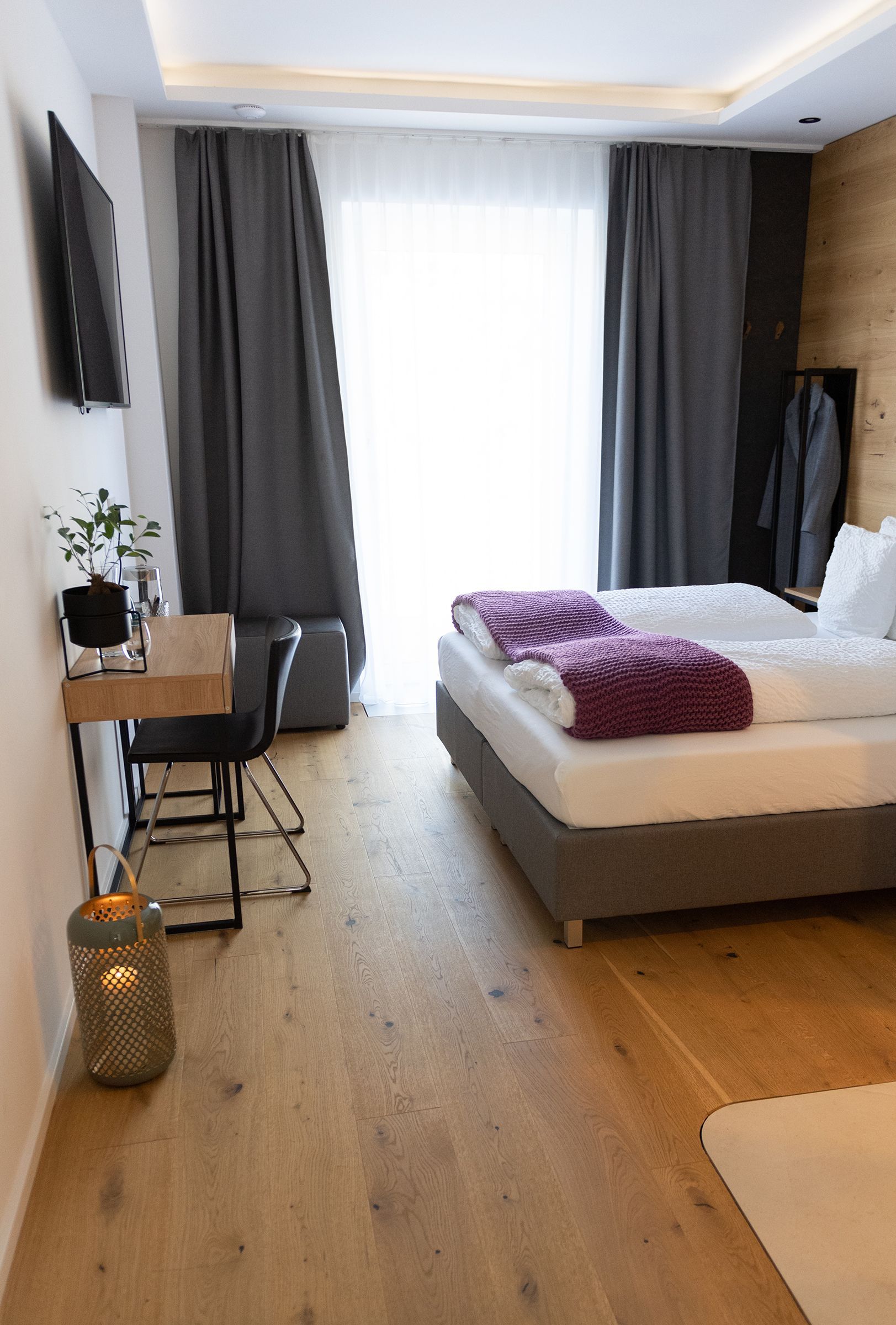 Das graue doppel-Boxspringbett mit weißen Laken steht im Fokus. Der Boden ist Parkett. Das Bett steht mit Kopfteil zu einer holzverkleideten Wand. Neben dem Bett ist eine große Fentstertüre mit Verdunklungsvorhängen. Gegenüber des Bettes ist ein Fernseher an der Wand und darunter ein schmaler Holztisch mit einem Stuhl.