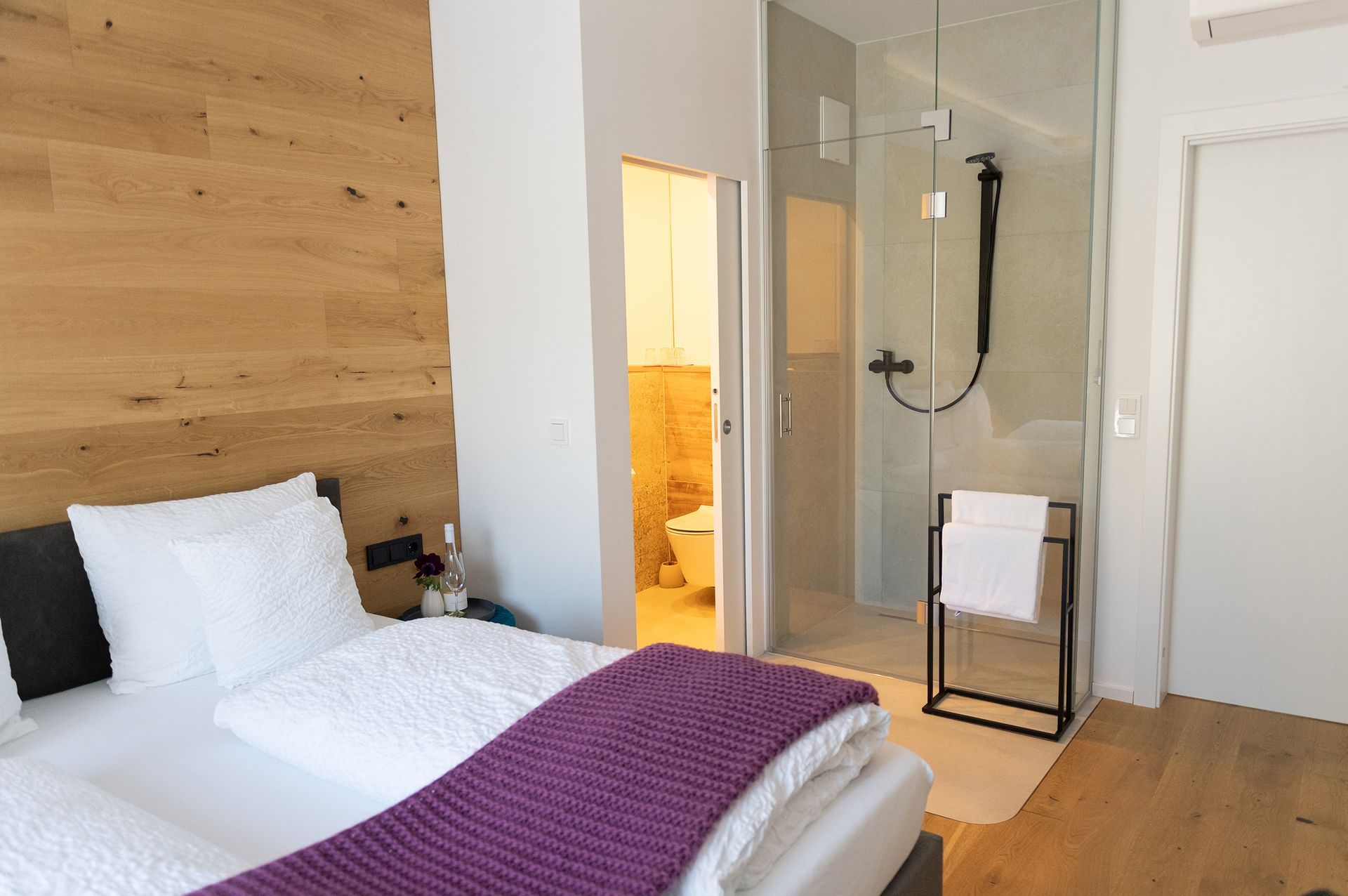 Ein Teil des grauen Bettes mit weißen Laken ist zu sehen. Daneben ist die moderne begehbare Dusche mit einer Glaswand abgetrennt. Neben dem Bett ist der Durchgang zum Badezimmer.