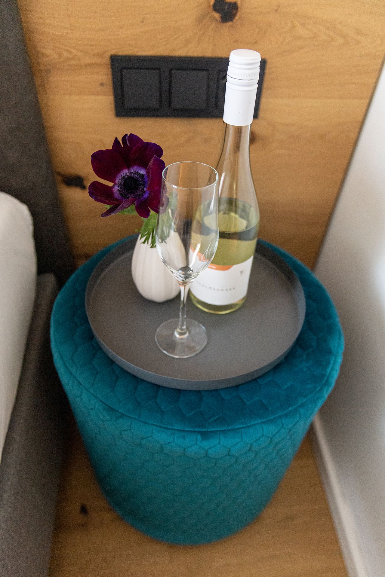 Türkiser Stoff-Beistellhocker neben Bett. Darauf ein schwarzes Tablett mit Weißweinflasche, Weinglas und lila Blumen in einer Vase. Dahinter schwarze Lichtschalter und eine holzverkleidete Wand.