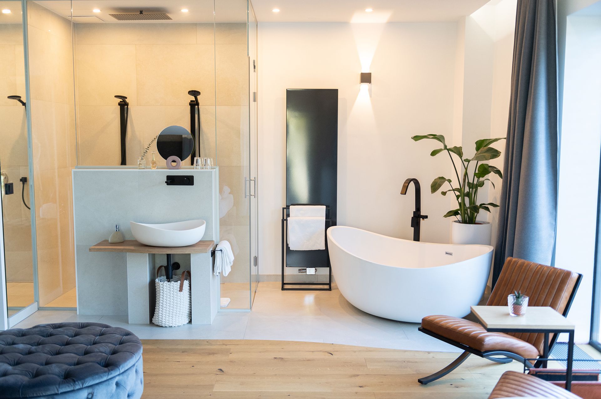 Offener Badezimmerbereich, freistehender Badewanne und begehbare Glasdusche. Eine große Pflanze steht hinter der Badewanne. An der Wand und in der Dusche ist eine warme, indirekte Beleuchtung.
