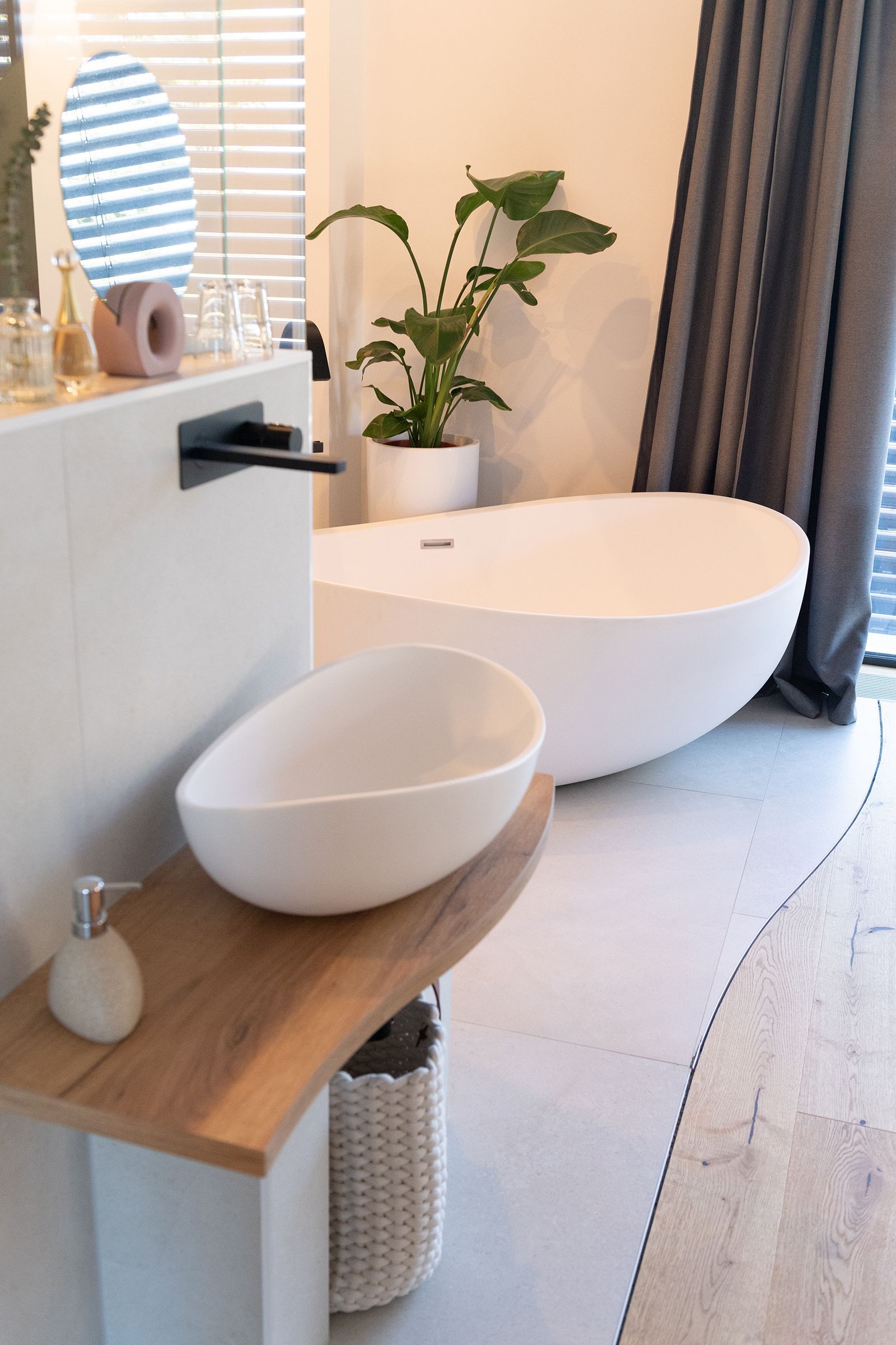 Offener Badezimmerbereich, freistehender Badewanne und weißes rundes Waschbecken. Eine große Pflanze steht hinter der Badewanne. Der Boden im Badezimmerbereich ist hellgrau verfliest. An der Wand ist eine warme, indirekte Beleuchtung.