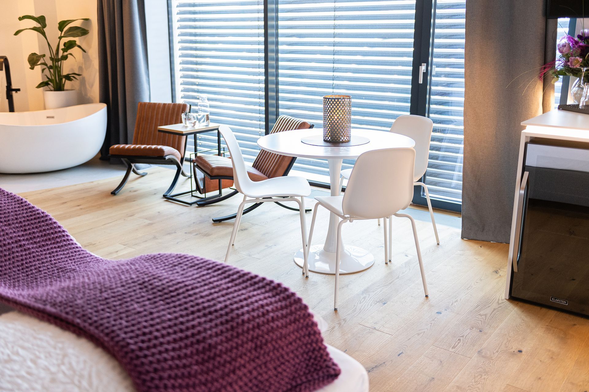 der weiße, runde Esstisch mit passenden Stühlen steht im Fokus. Dahinter zwei braune Lederstühle mit schwarzem Gestell aus Metall stehen an der Fensterwand und Rollläden. Dazwischen ein kleiner Beistelltisch. 