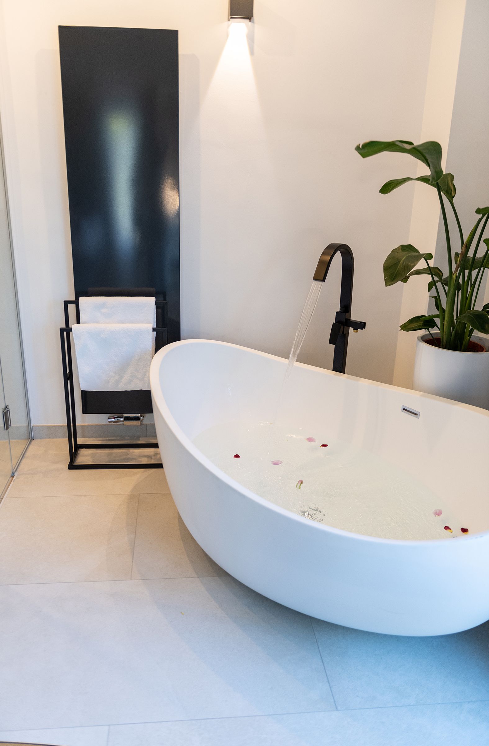 Offener Badezimmerbereich, freistehender Badewanne mit schwarzen Armaturen. Eine große Pflanze steht hinter der Badewanne. Der Boden im Badezimmerbereich ist hellgrau verfliest. An der Wand  ist eine warme, indirekte Beleuchtung.