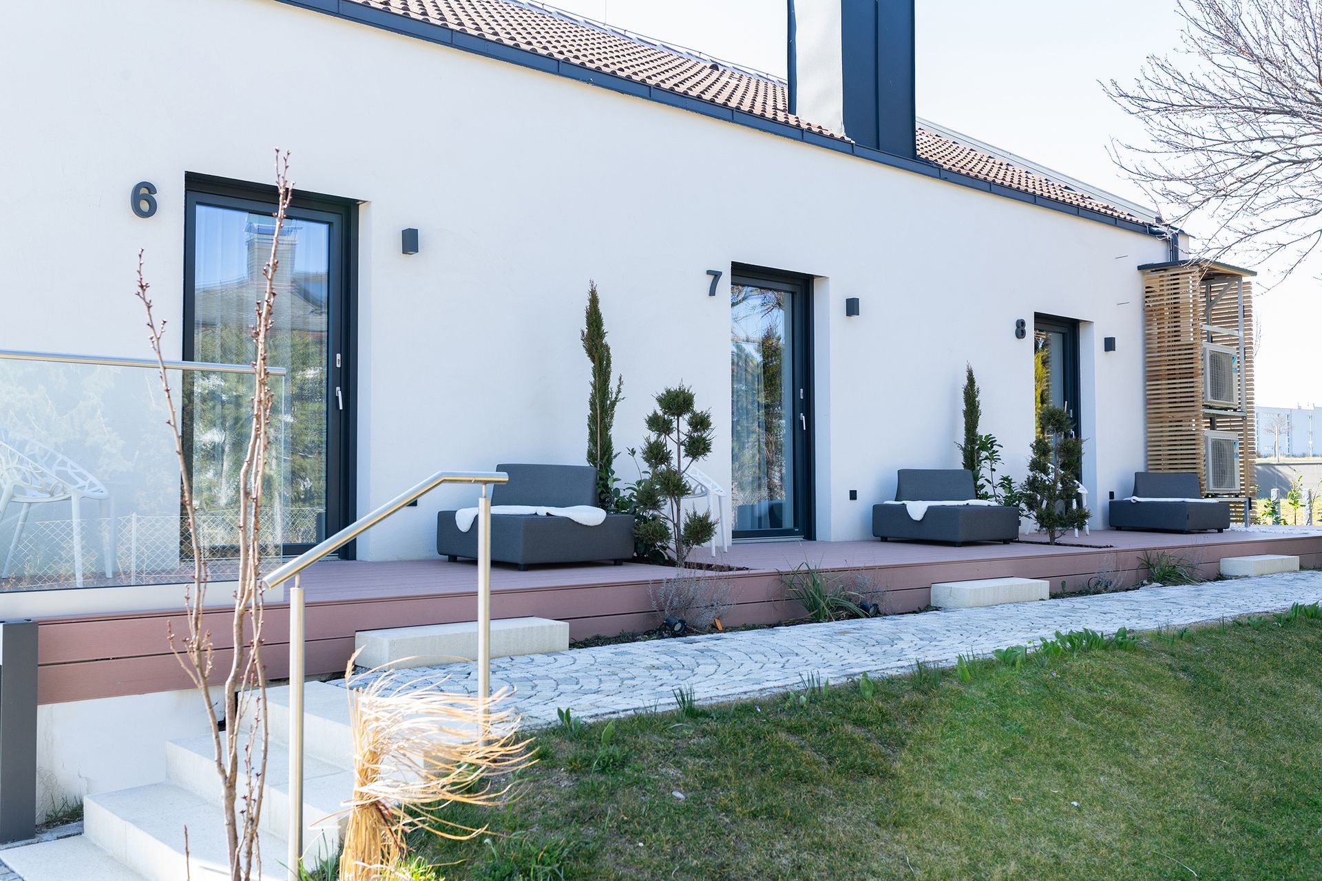 Außenansicht der Holzterrassen von Apartment 6, 7 und 8. Jede Terrasse hat einen grauen Stoffsessel und Pflanzen. Die weiße Wand und die grauen Fenstertüren bilden einen Kontrast. 