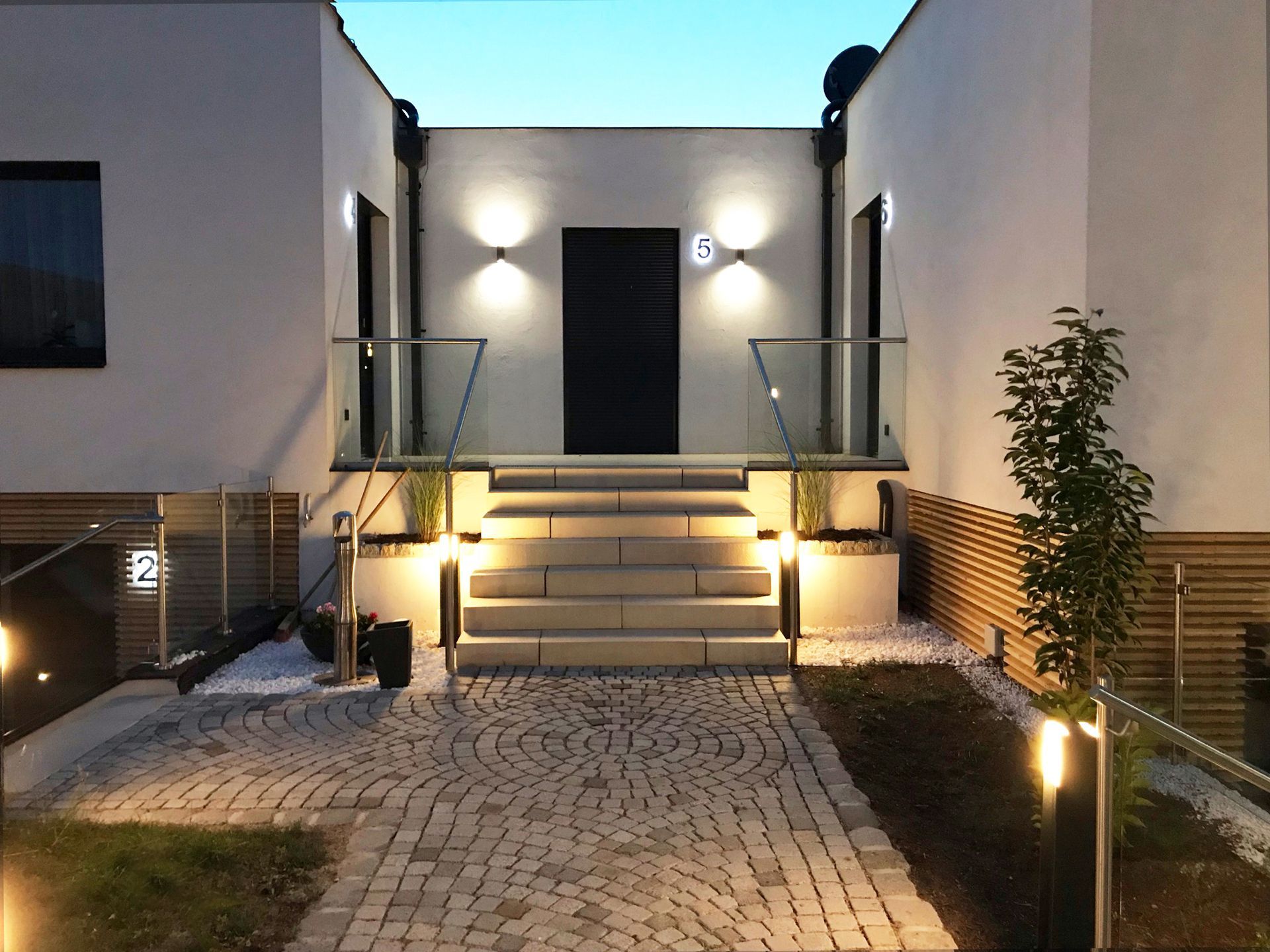 Außenansicht von Verbindungs-Haus der Geisler Appartments, graue Stufen führen zum Eingang. Der Pflastersteinweg führt zu den Stufen. Der Weg ist bei Dämmerung beleuchtet.