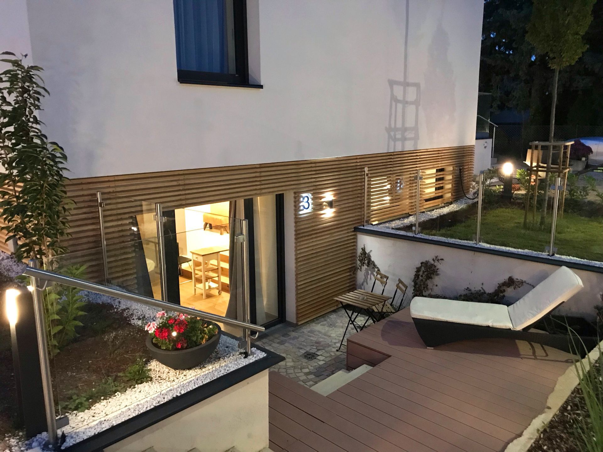 Außenbereich mit Holzterrasse, einem Holztisch sowie zwei passende Stühle. Indirekte Beleuchtung findet sich in der Holzfassade an der Wand des Eingangs zu Apartment 3.