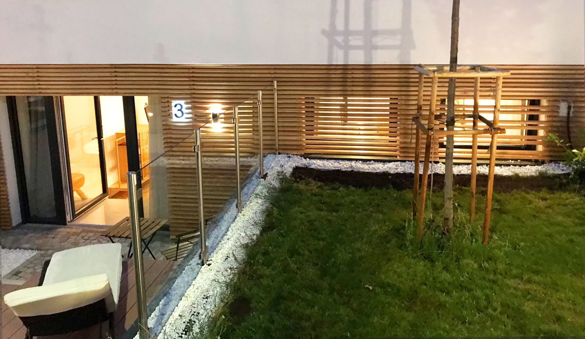 Außenansicht der erhöhten Rasenfläche und Glasgeländer. Indirekte Beleuchtung findet sich in der Holzfassade an der Wand des Eingangs zu Apartment 3.
