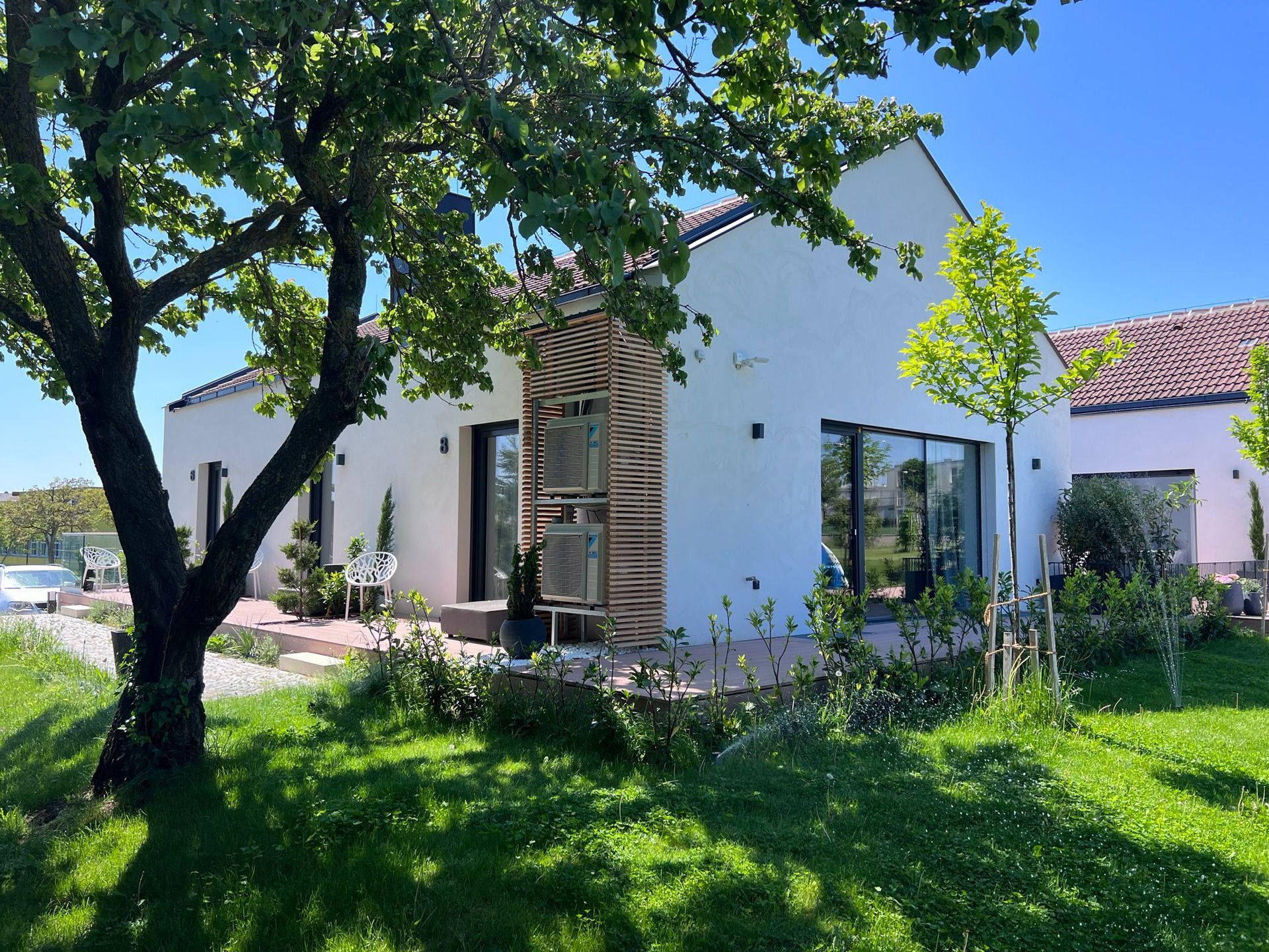 Außenansicht der Holzterrassen von Apartment 6, 7 und 8. Jede Terrasse hat einen grauen Stoffsessel und Pflanzen. Der Rasen ist gepflegt und grün. Ein Baum ragt von der Seite in das Bild hinein.