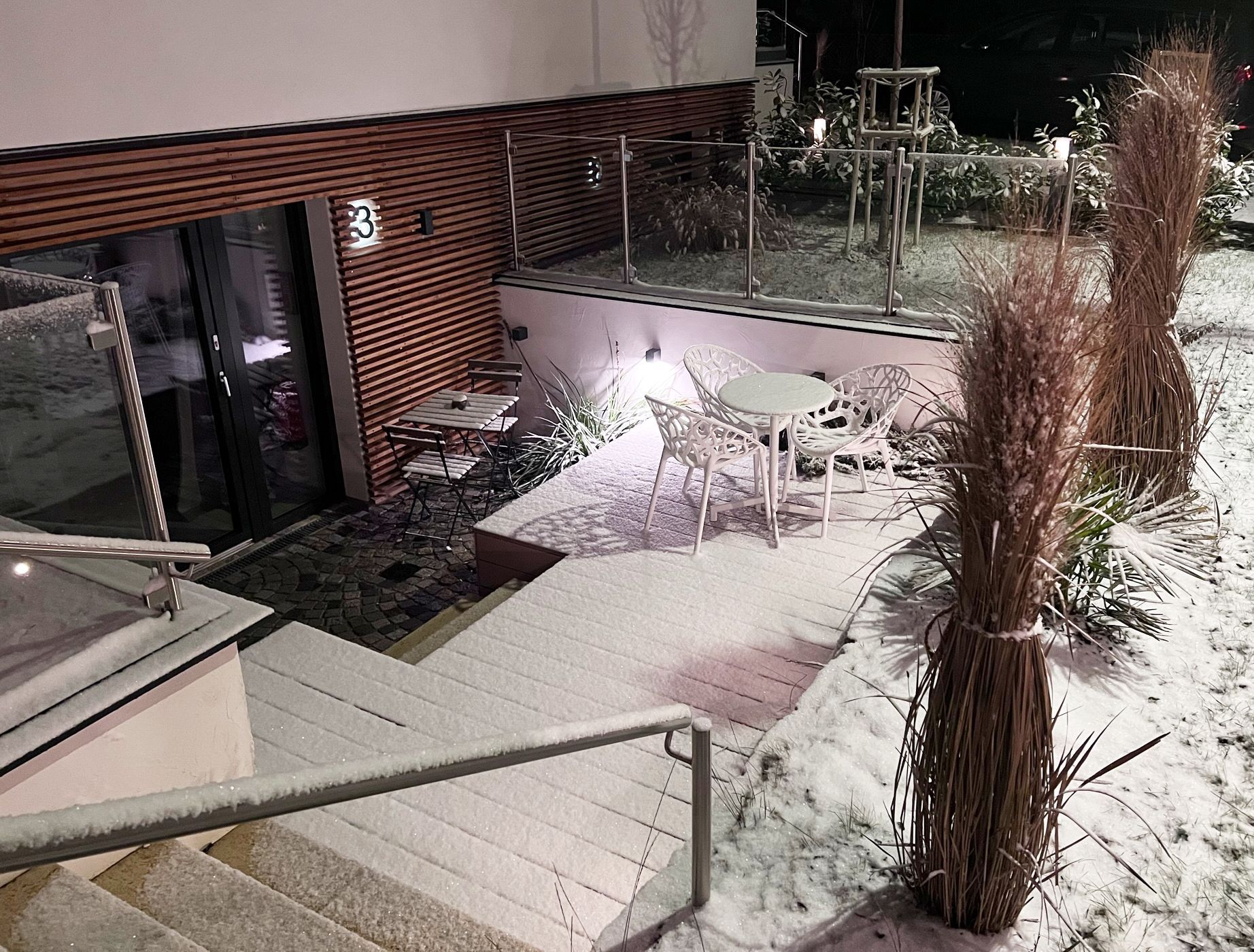 Außenbereich mit Holzterrasse, einem Holztisch sowie zwei passende Stühle. Indirekte Beleuchtung findet sich in der Holzfassade an der Wand des Eingangs zu Apartment 3. Boden und angrenzende Stiegen sind mit Schnee bedeckt.