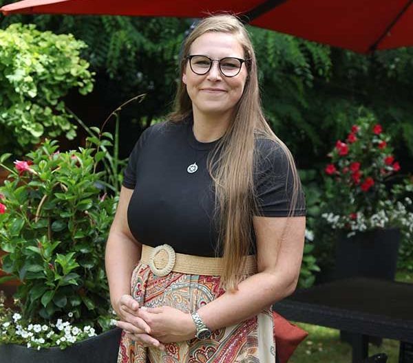 Eine Person mit langen Haaren und Brille trägt ein schwarzes Hemd und einen gemusterten Rock und lächelt, während sie in einem Garten steht.