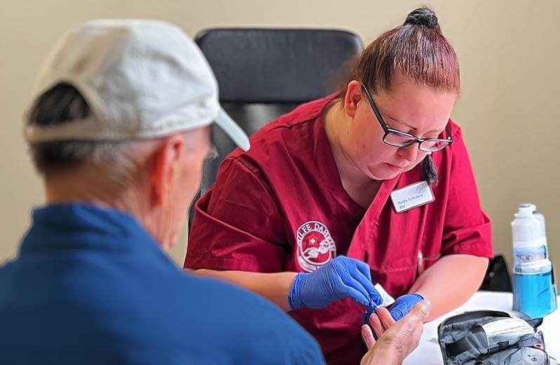 Eine medizinische Fachkraft in weinroter Uniform und blauen Handschuhen führt bei einem Patienten eine Blutzuckermessung mittels Fingerstich durch.
