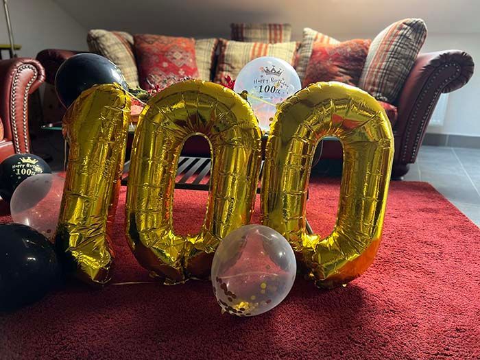 Große goldene Folienballons mit der Aufschrift „100“ stehen auf einem roten Teppich vor einem Sofa, umgeben von mehreren kleineren, durchscheinenden Ballons.
