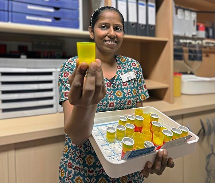 Eine Mitarbeiterin des Gesundheitswesens in gemusterter Uniform hält einen kleinen gelben Medikamentenbecher hoch und trägt ein Tablett mit anderen Bechern in einer Klinik.