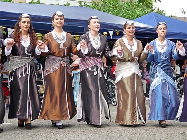 Fünf Personen in traditionellen Seidengewändern und Kopfbedeckungen tanzen im Freien unter einem blauen Baldachin in einer Reihe.